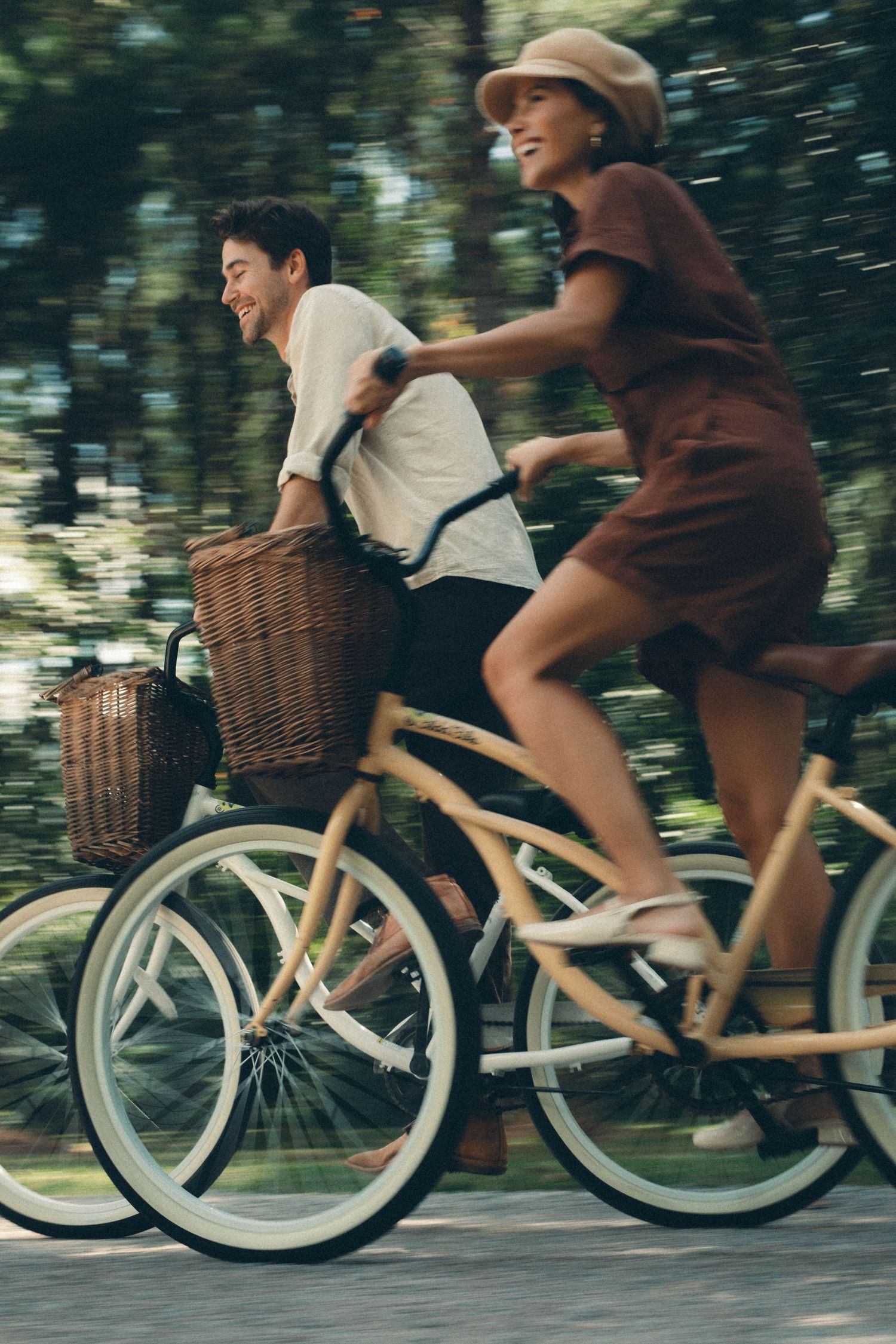 Two people ride vintage-style bicycles with wicker baskets through a tree-lined path in motion blur.