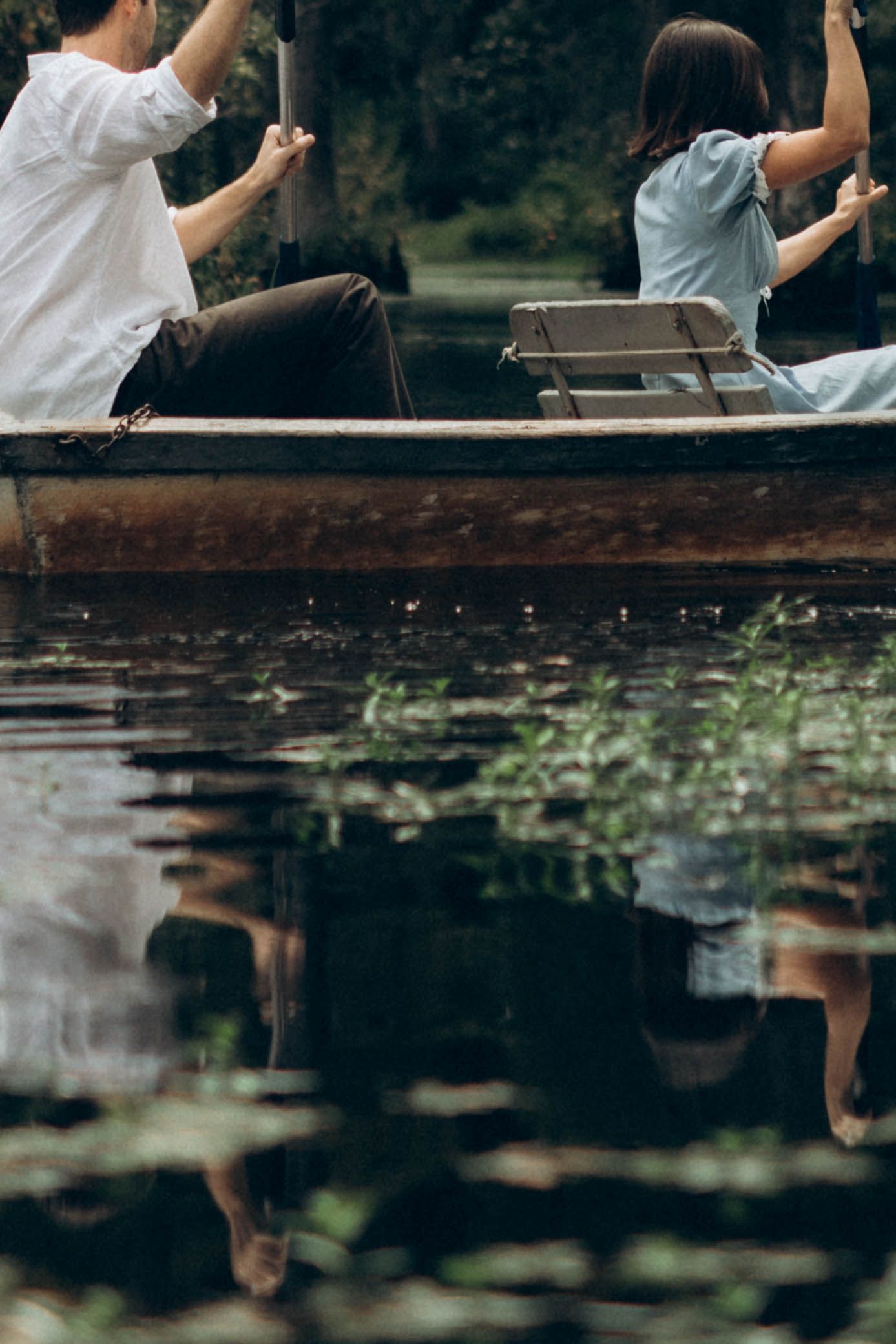 A couple sitting in a wooden rowboat on a tranquil lake surrounded by greenery.