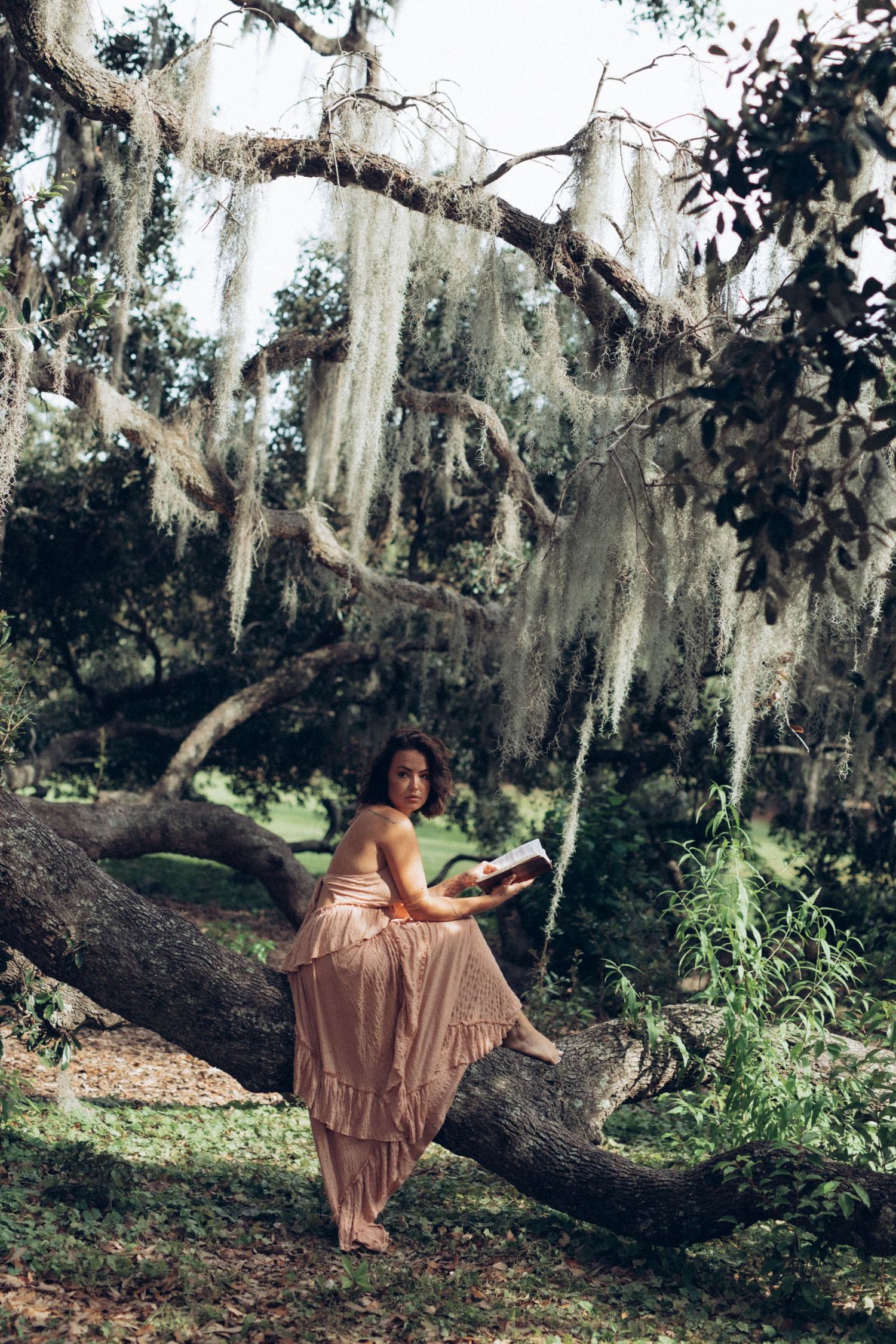 Person in flowy dress poses gracefully on tree branches draped with Spanish moss in enchanted forest setting.
