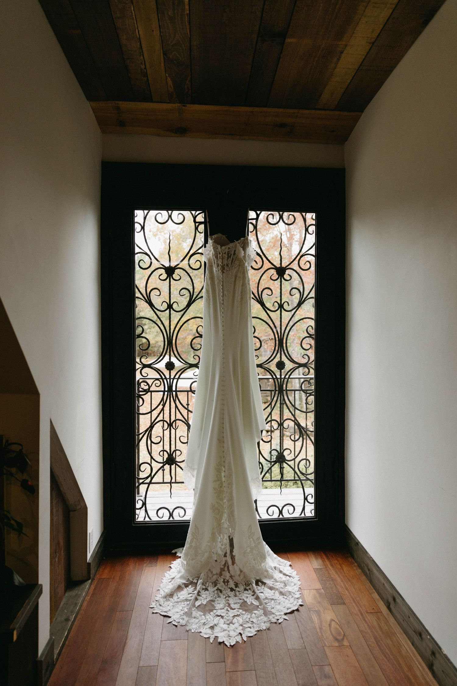 A white wedding dress hangs elegantly in front of ornate wrought iron windows in a rustic room with wooden floors and beams.