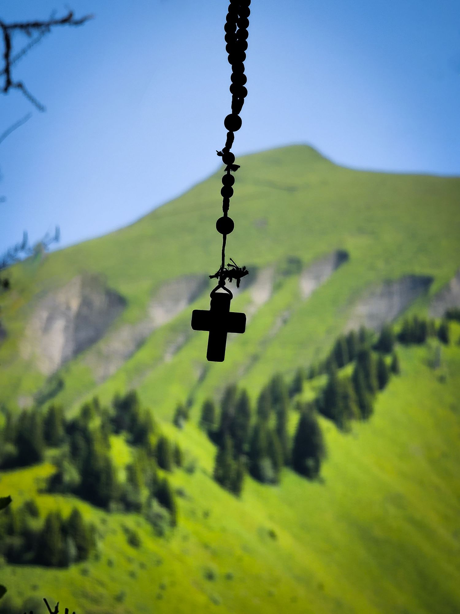 Un chapelet en silhouette avec une croix est suspendu sur un fond de pentes de montagne verdoyantes sous un ciel bleu.