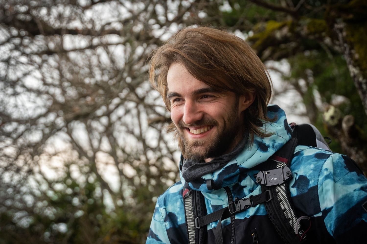 Solo hiker in blue camo jacket smiles while enjoying outdoor adventure on wooded trail.