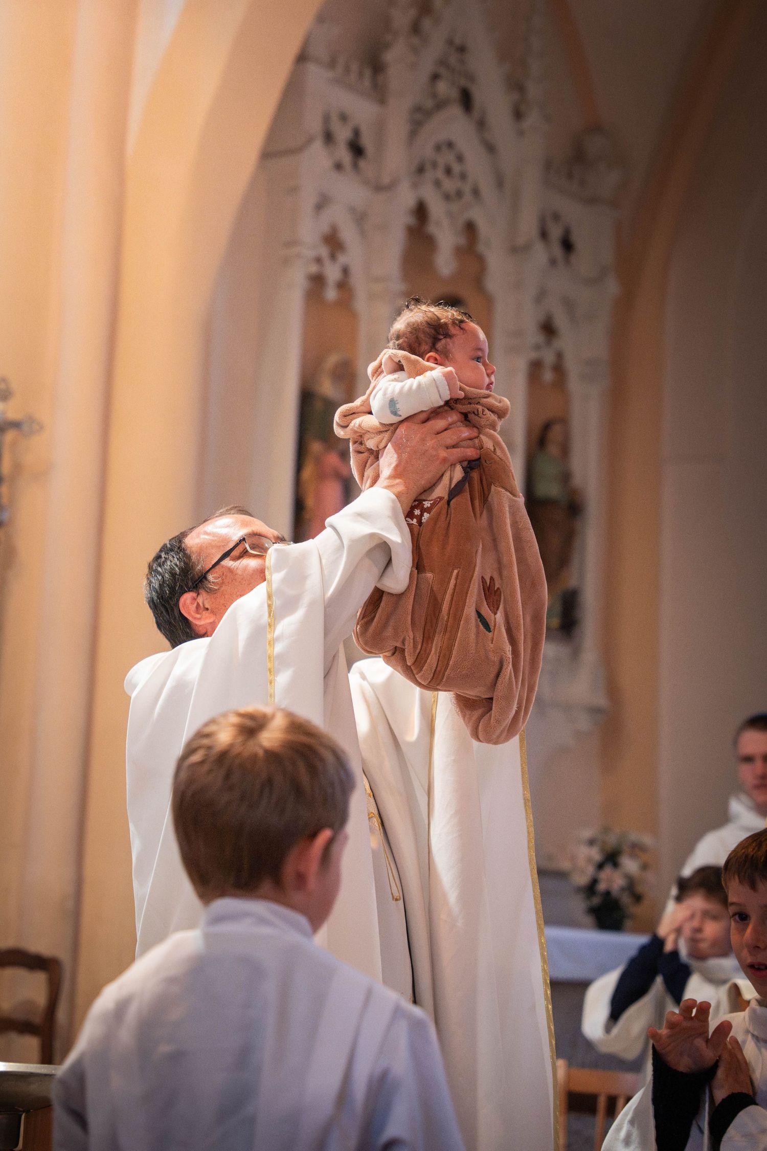 Priest lifting baby during baptism ceremony in a church, with children and congregation watching in the background.