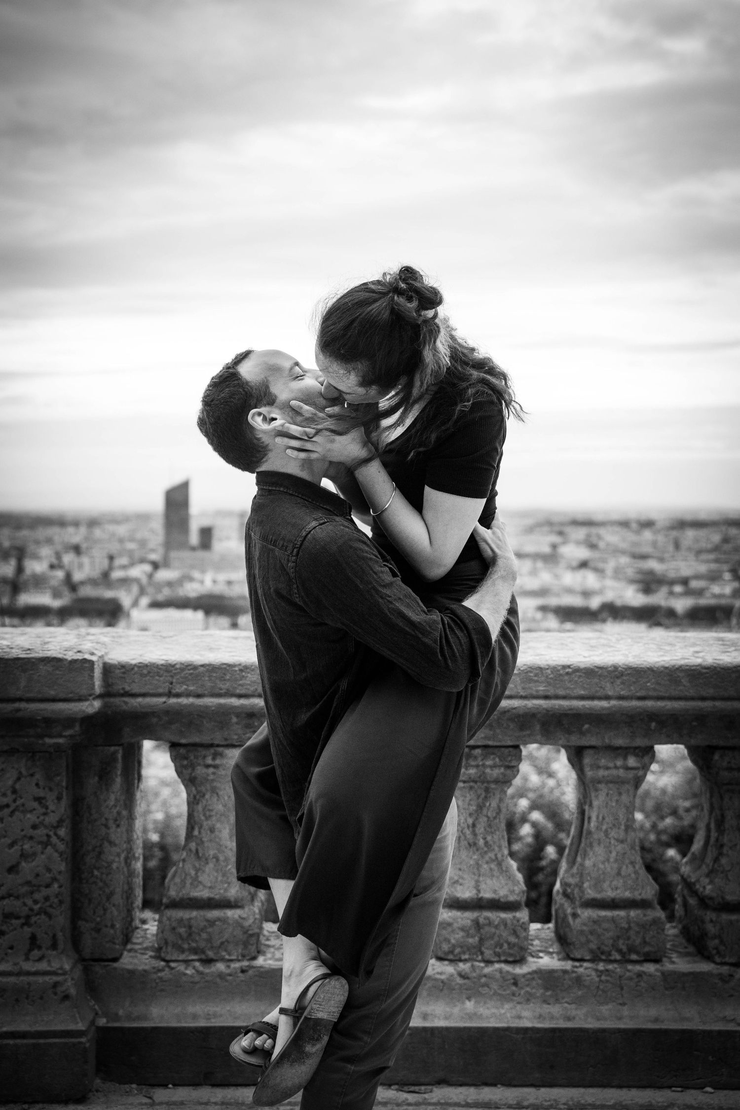 Un couple romantique partageant un baiser passionné sur un balcon surplombant le paysage urbain de Lyon en photographie noir et blanc.