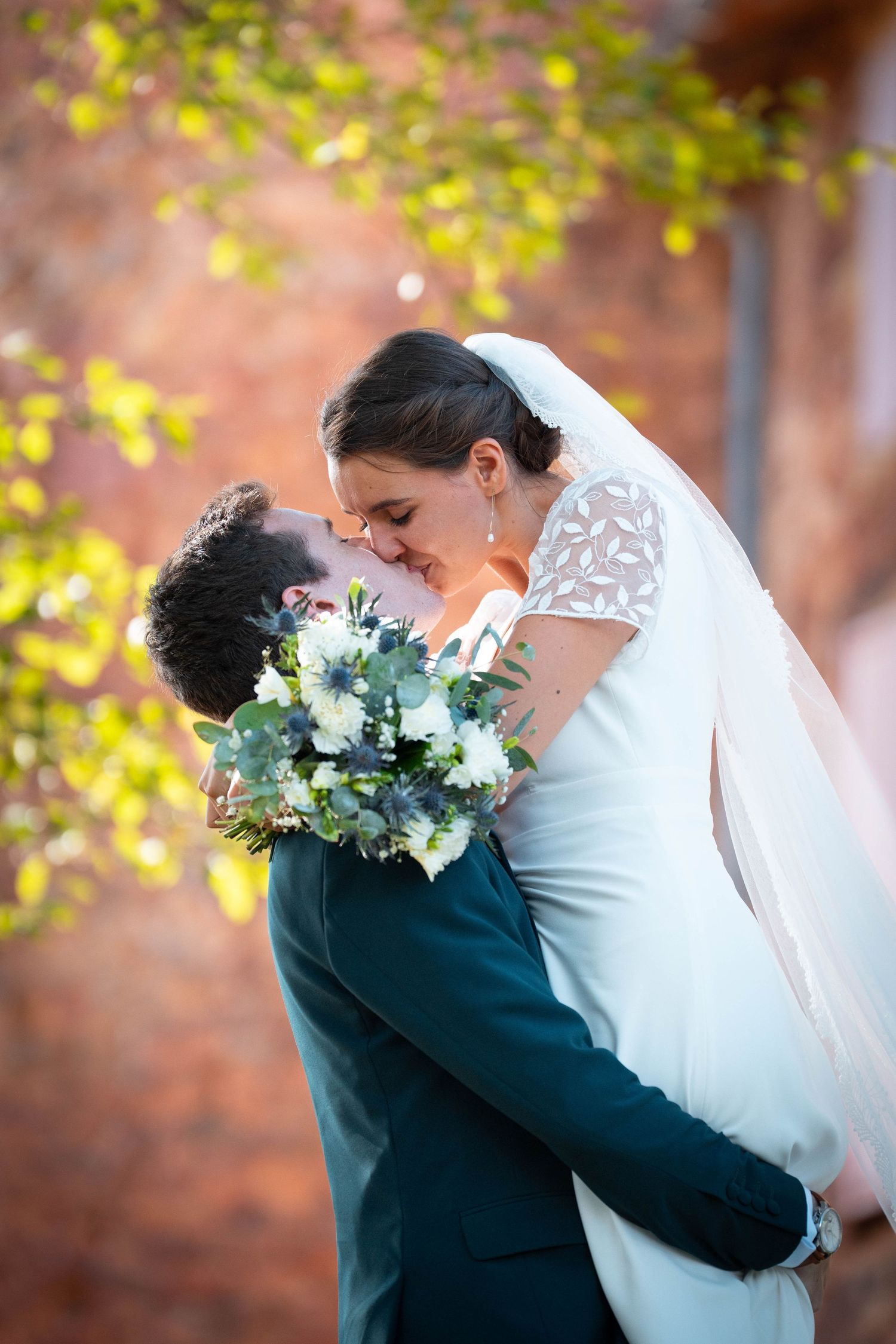 Bride and groom sharing a romantic kiss, bride in white gown holding bouquet, groom in teal suit, autumn backdrop.