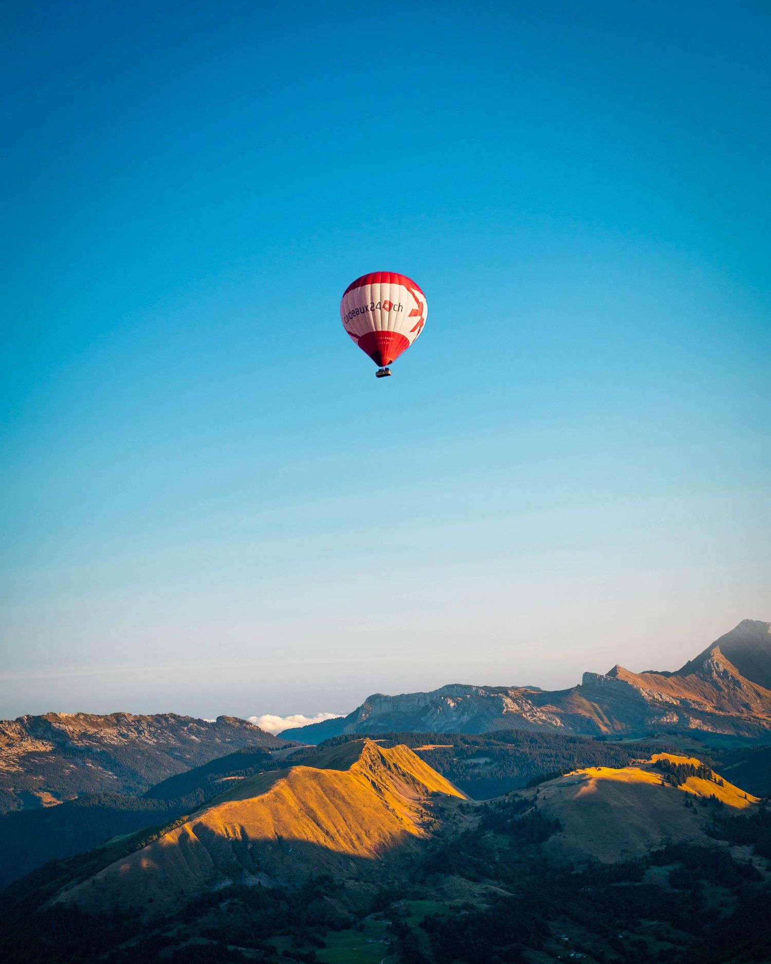 Une montgolfière rouge flotte au-dessus des sommets dorés des montagnes contre un ciel bleu vibrant au lever du soleil.