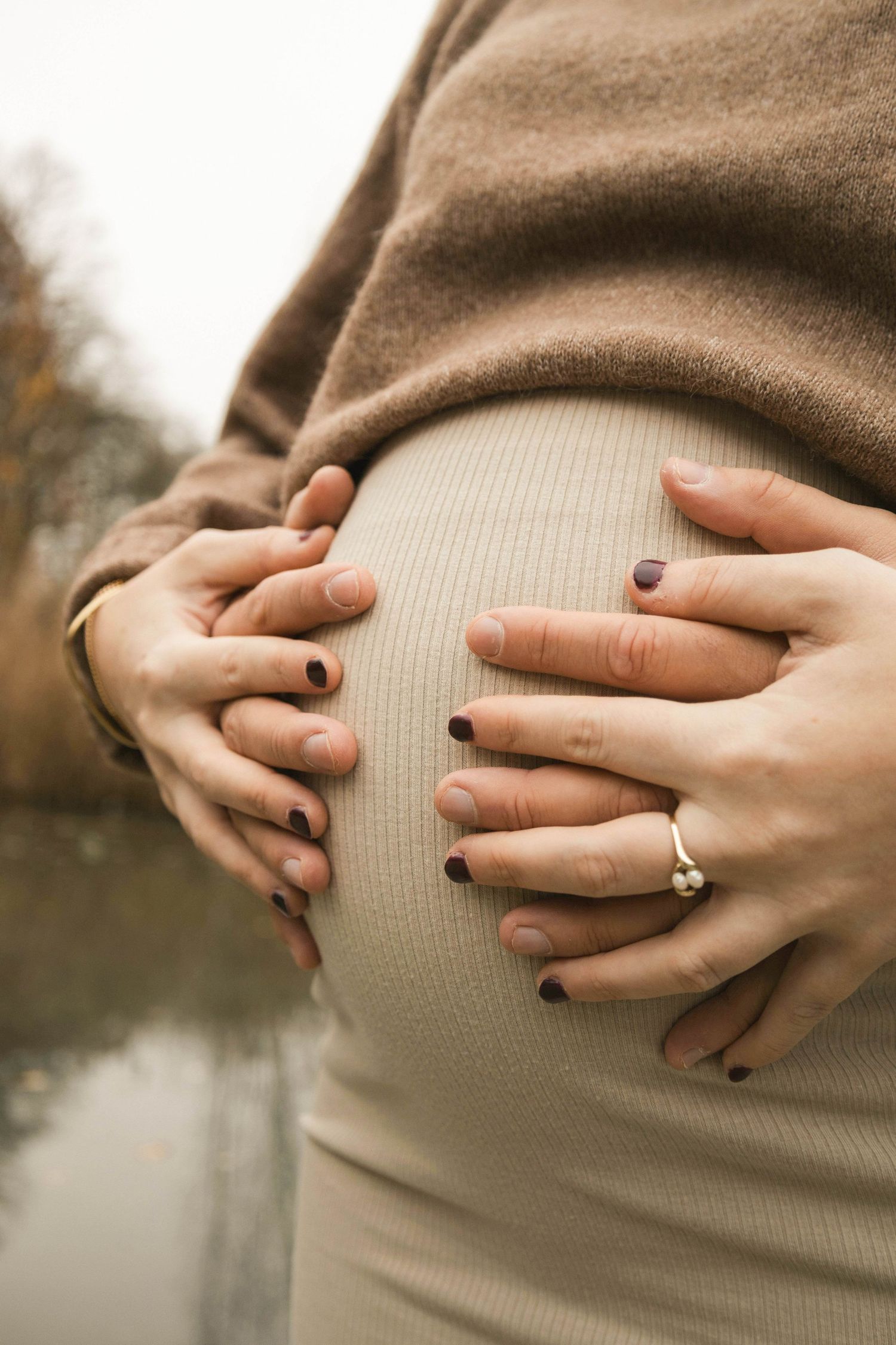 Pregnant woman in beige outfit cradling baby bump with both hands showing dark nail polish and diamond ring outdoors.