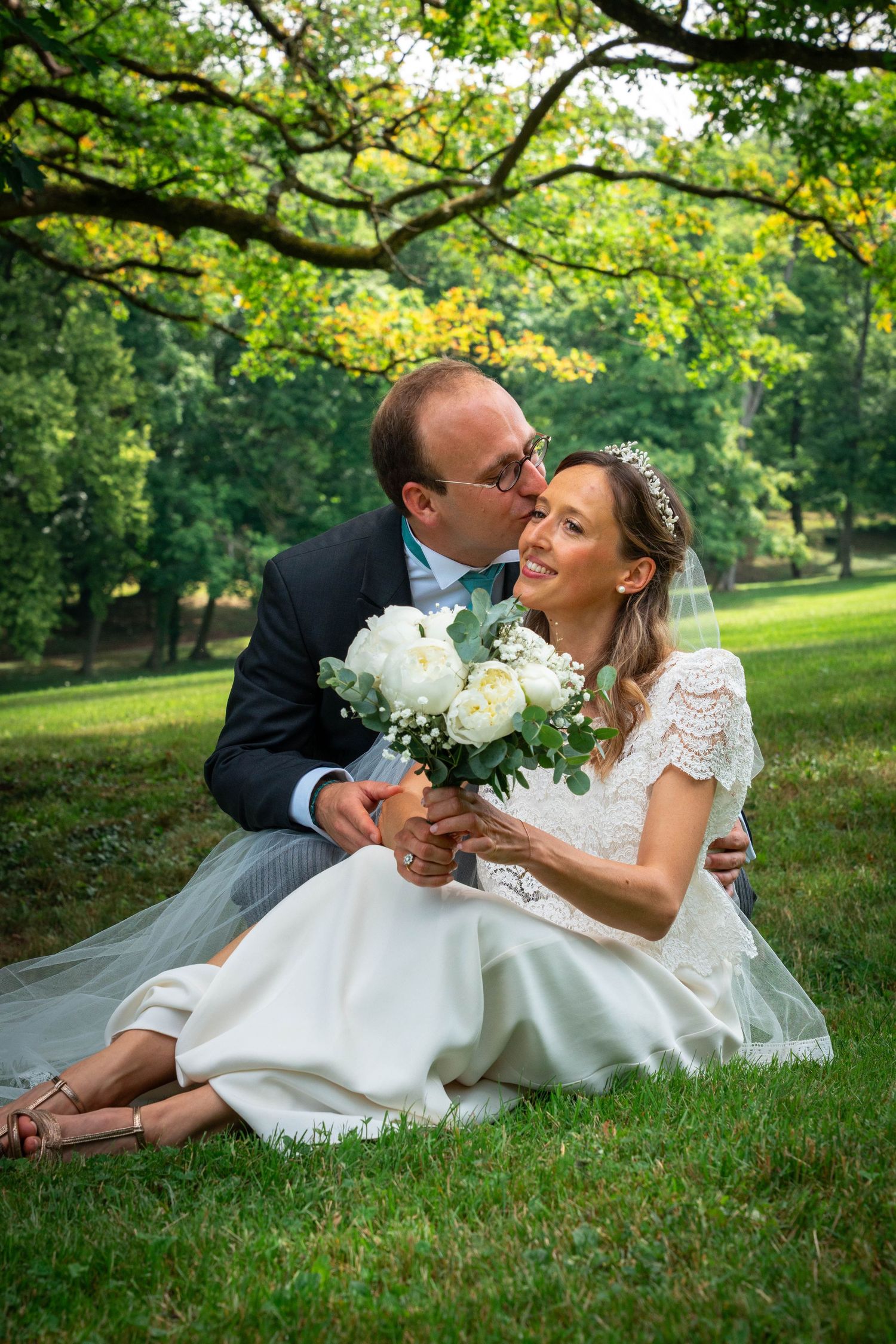 A bride and groom sit on green grass under a tree, sharing a tender moment with a white bouquet.