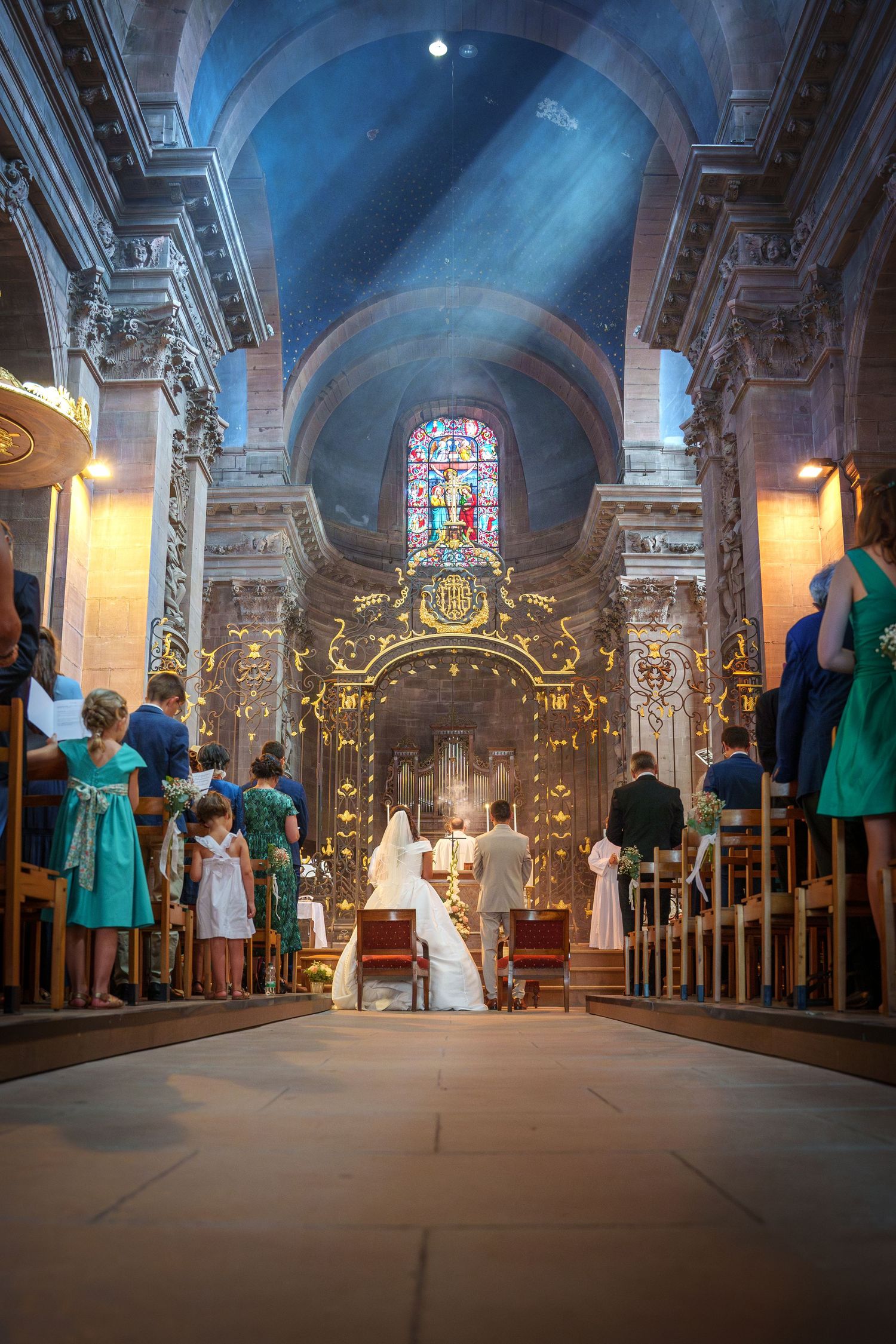 Des faisceaux lumineux traversent les vitraux d'une cathédrale lors d'une cérémonie de mariage avec des participants vêtus de turquoise.