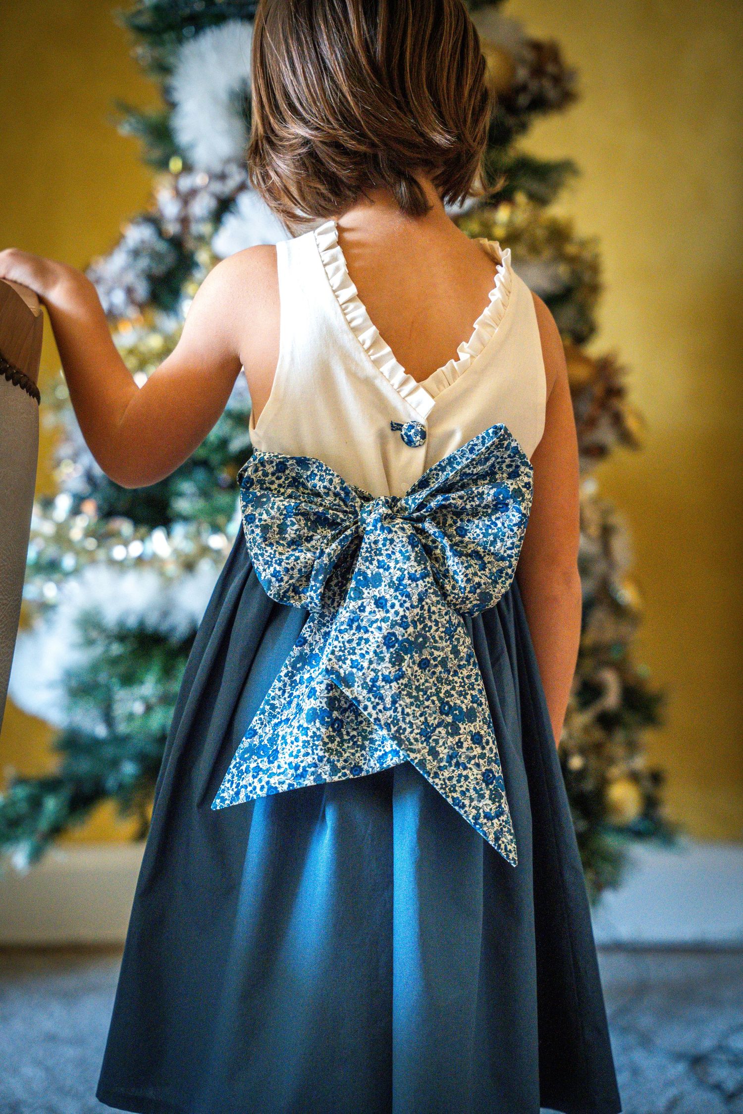 Vue arrière d'une élégante robe bleu marine avec des bretelles à volants blancs et un grand nœud floral sur fond d'arbre de Noël.