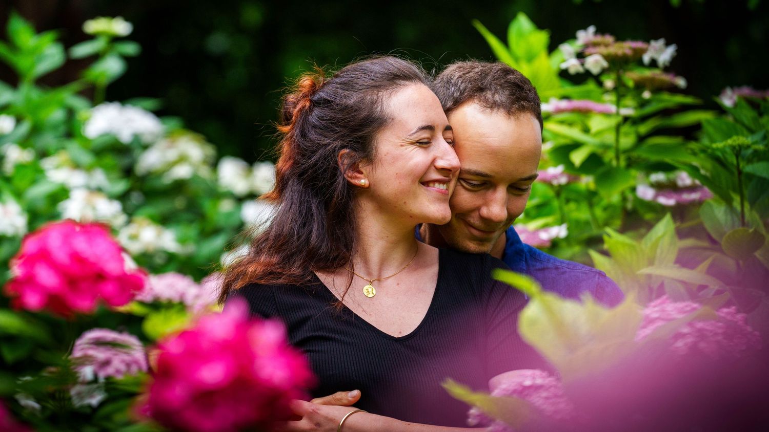 Couple smiling and embracing in a lush garden surrounded by vibrant pink and white flowers.