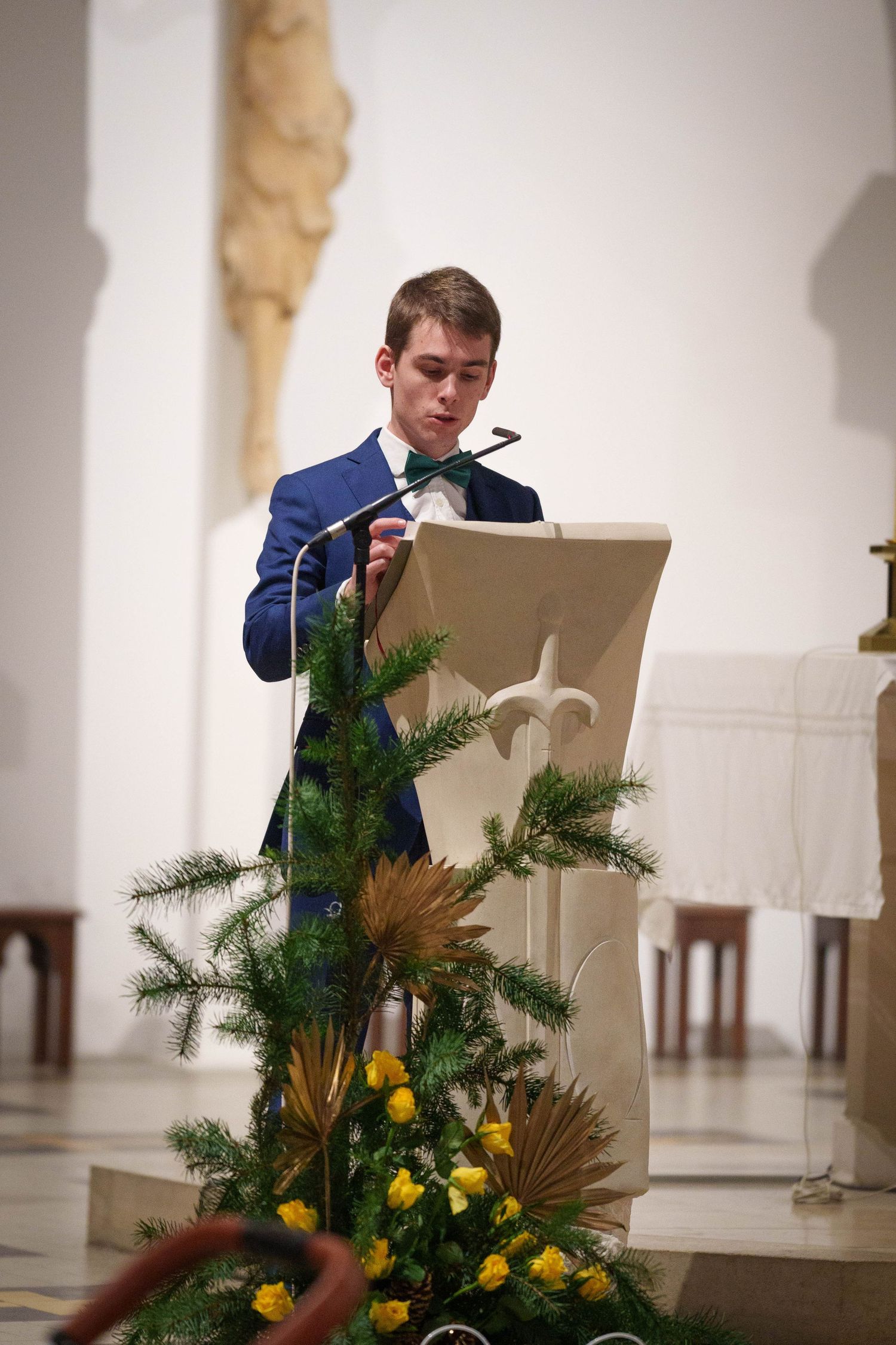 Une personne en costume bleu marine lit sur un pupitre décoré de fleurs jaunes et de branches de pins dans un décor d'église lumineuse.
