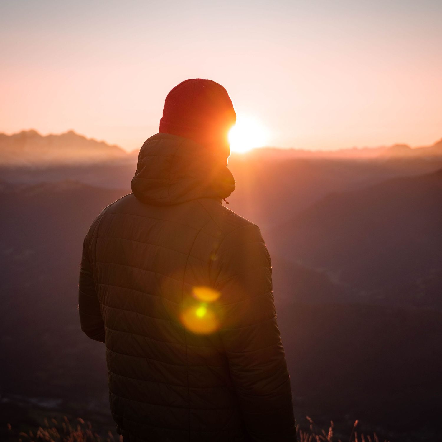 Une silhouette se dresse devant un coucher de soleil majestueux sur les sommets des montagnes avec un éclat de soleil doré en arrière-plan.