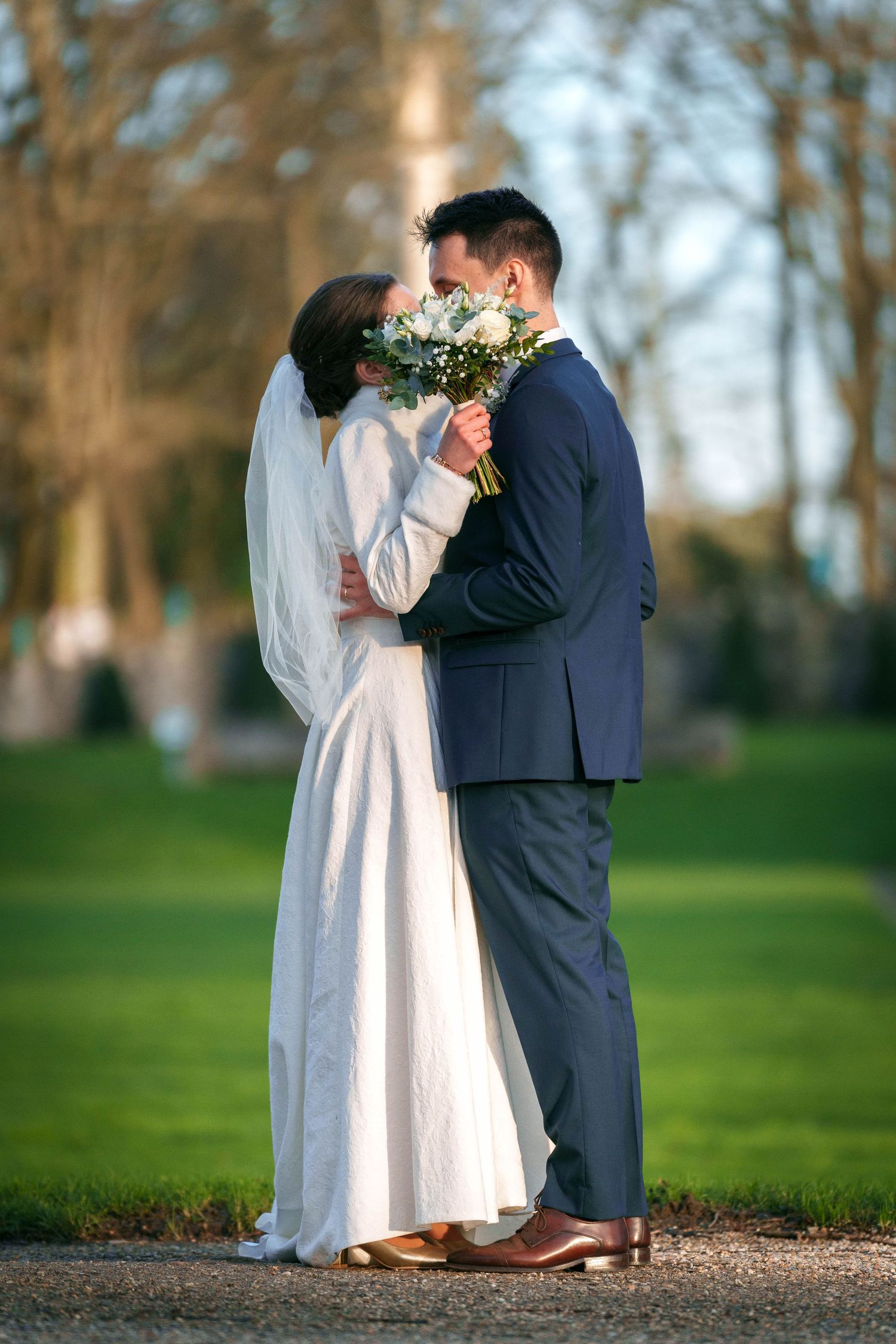 Romantic newlyweds share a tender moment on a garden path while holding a white floral bouquet.