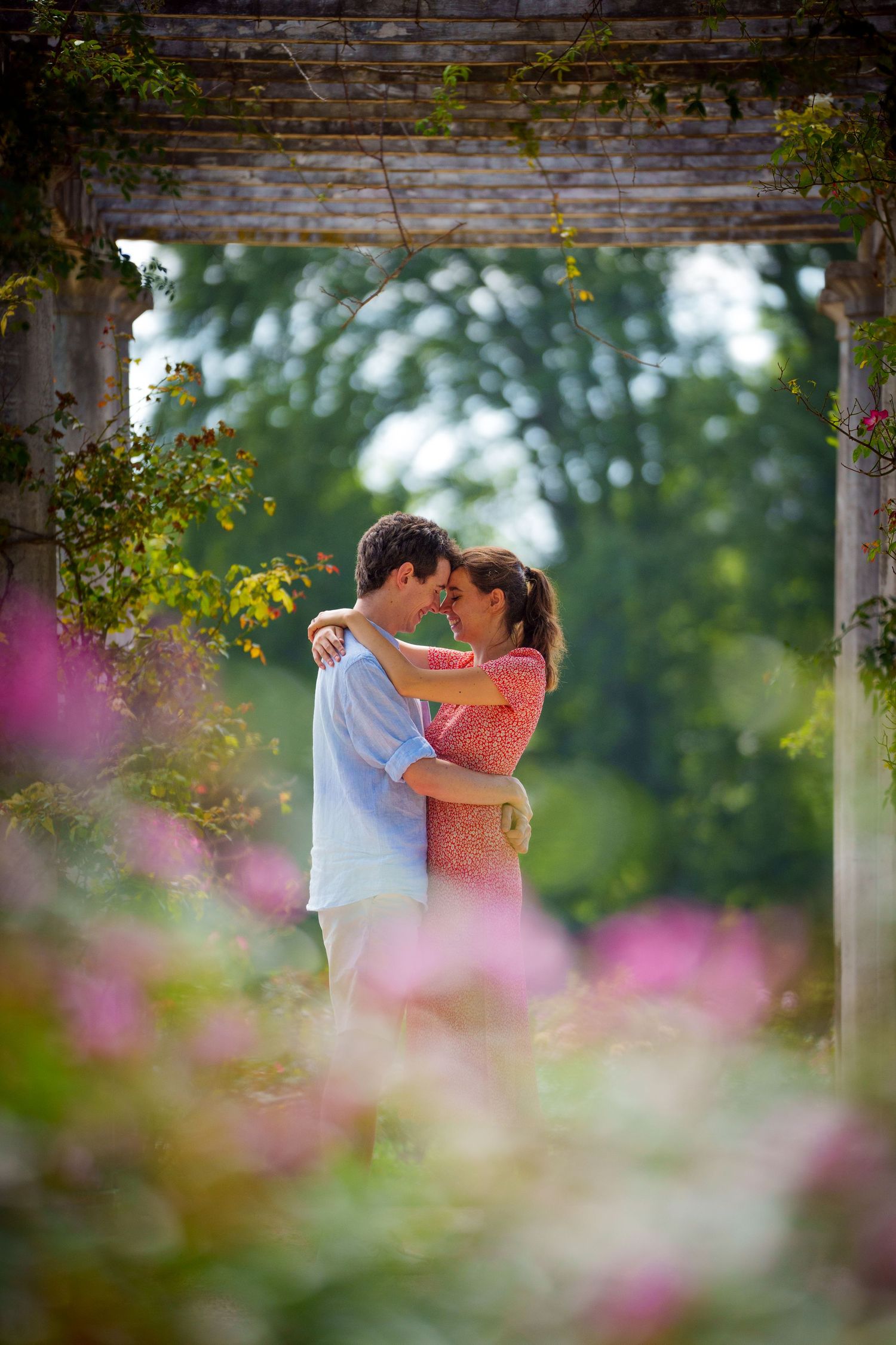 Couple embracing tenderly under a rustic pergola surrounded by blooming pink flowers in a lush garden.