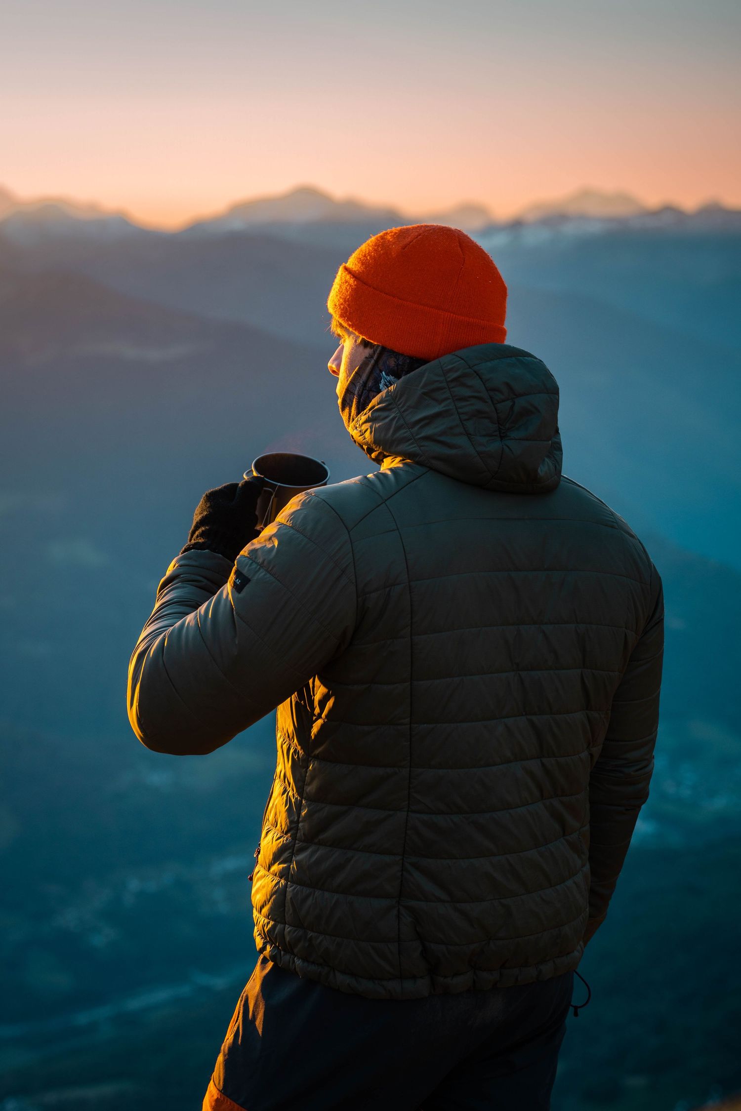 Une personne portant une veste bouffante et un bonnet orange regarde les sommets des montagnes pendant l'heure dorée du coucher du soleil.