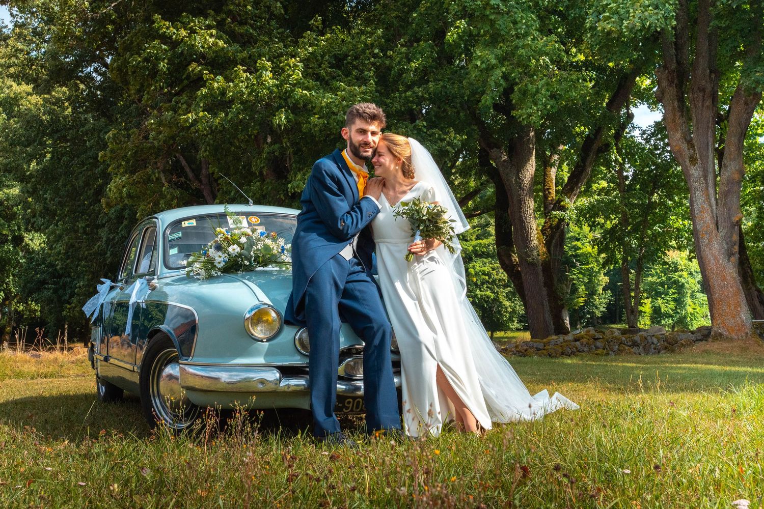Bride and groom pose beside a vintage light blue car decorated with flowers in a lush green park setting.