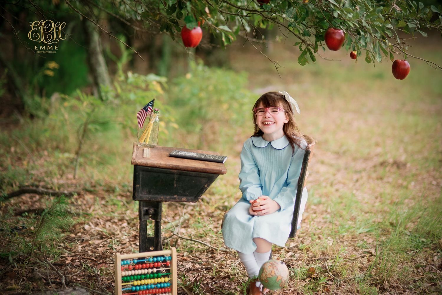 Young girl in blue dress sits at vintage school desk under apple tree in a charming outdoor back-to-school photo session in Orangeburg, SC