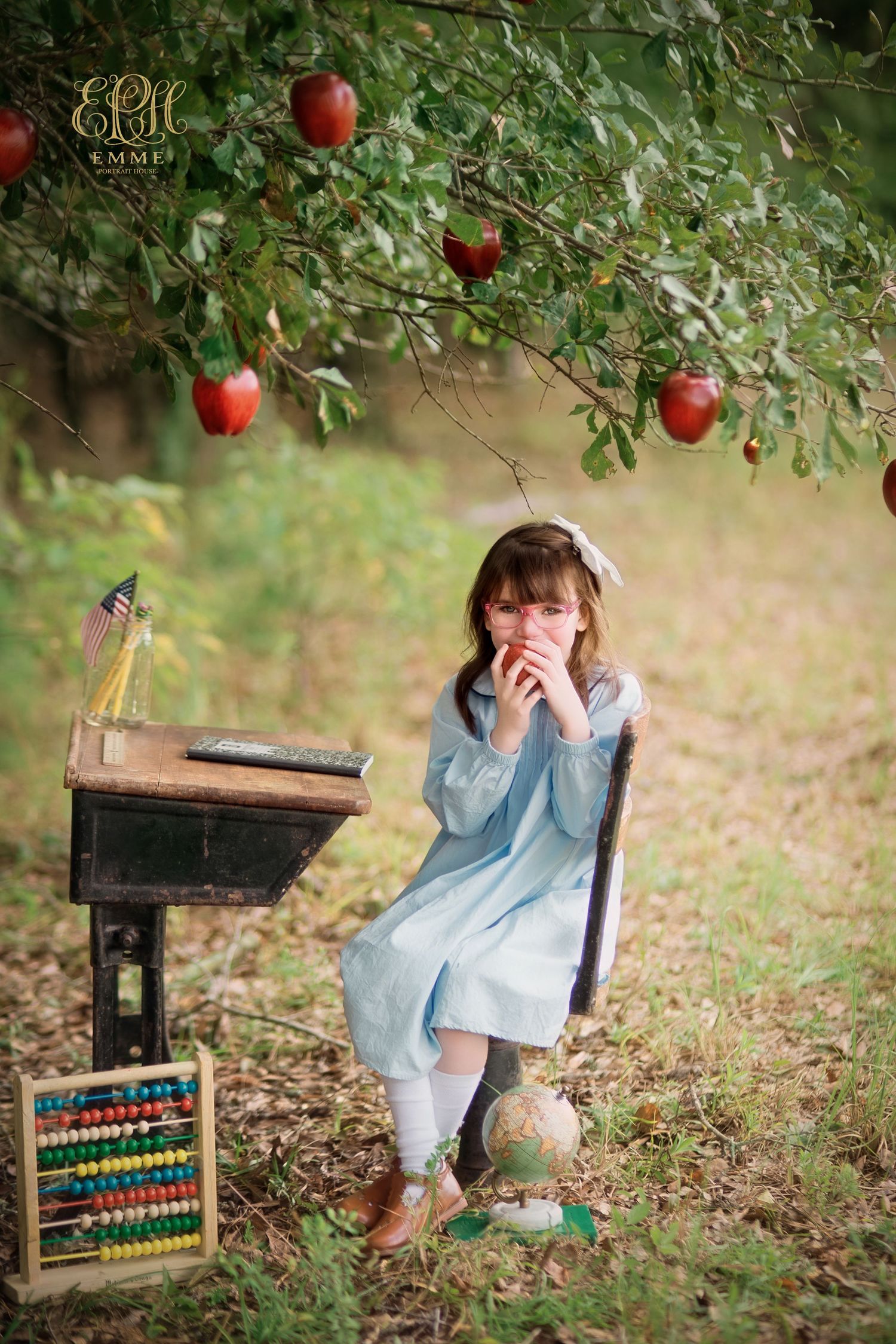 A young girl in a blue dress sits under an apple tree with a vintage desk, globe, and abacus in a garden setting in Orangeburg, SC