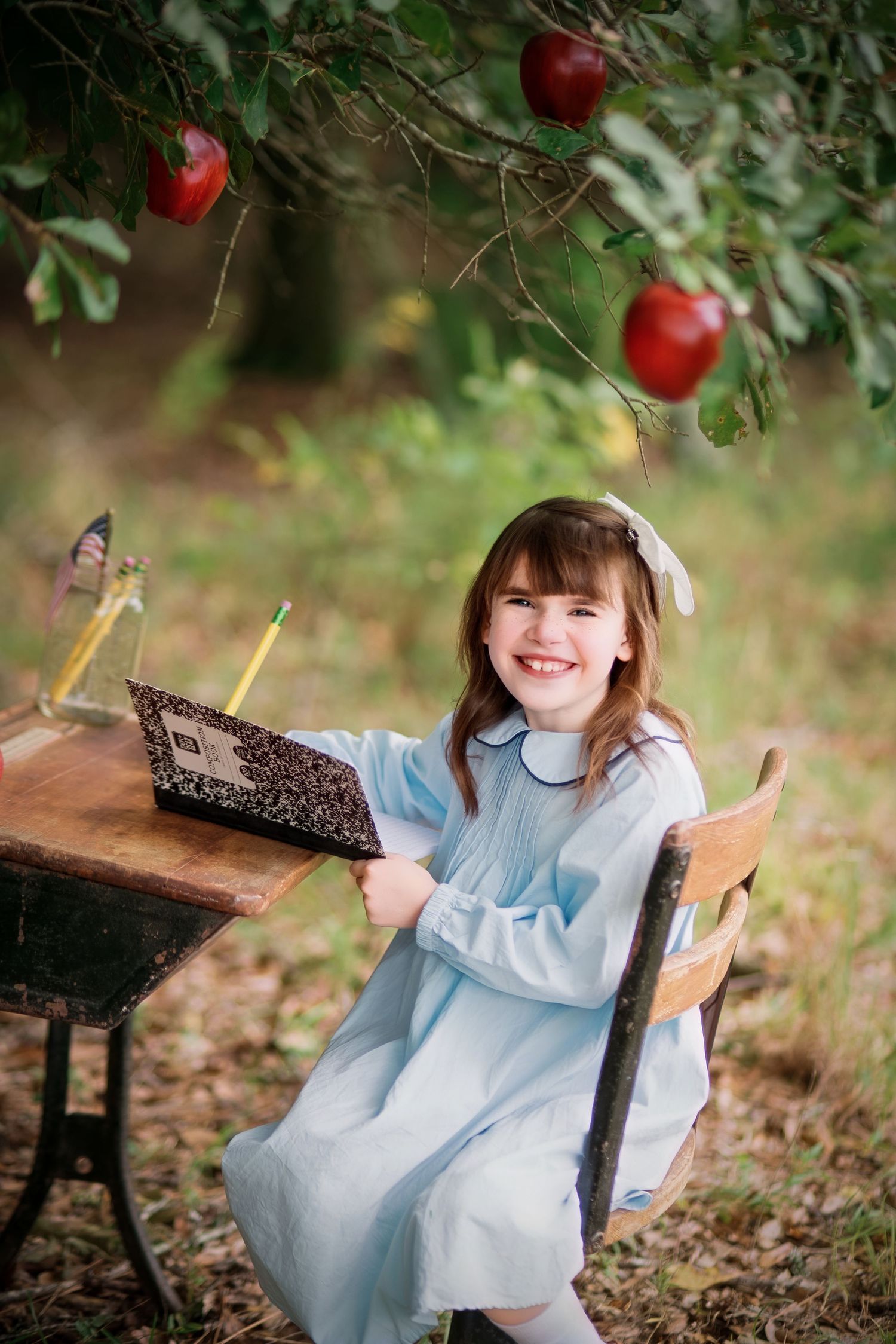 A smiling girl in a blue dress sits at a wooden desk under an apple tree, holding a composition notebook outdoors in Orangeburg, SC