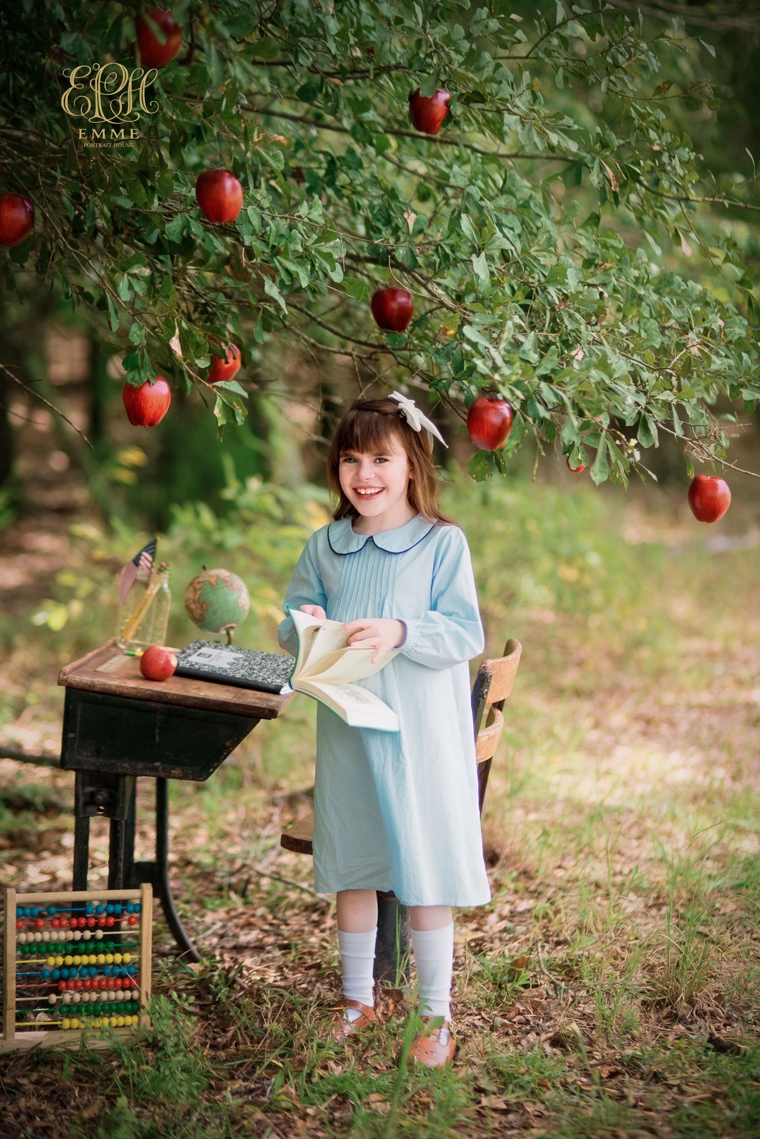 Young girl in blue dress holding book under apple tree with vintage school desk, globe, and abacus outdoors in Orangeburg, SC