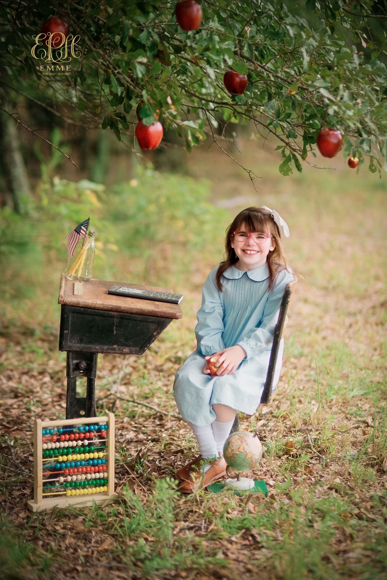 A smiling girl in a blue dress sits outdoors by a school desk with an abacus and globe under an apple tree in Orangeburg, SC