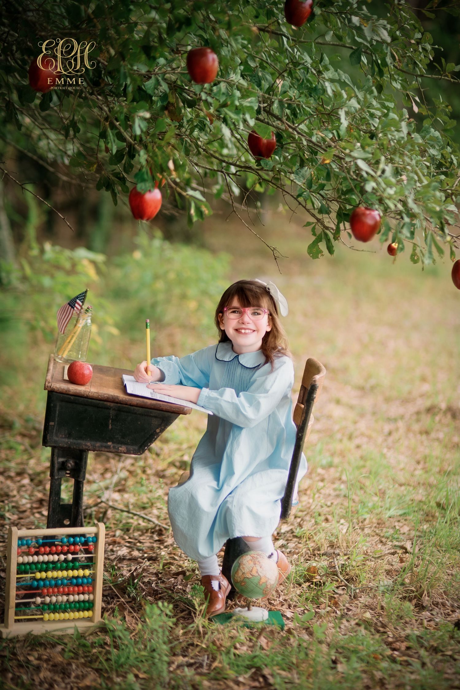 Girl in blue dress sits at vintage school desk under apple tree with abacus and globe in a charming outdoor setting in Orangeburg, SC