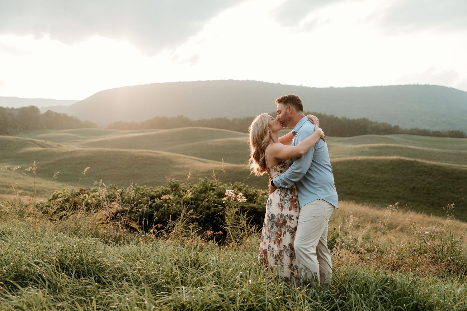 couple embracing and kissing while the sun sets on the mountain range behind them. Soft sun rays fall acoss the land