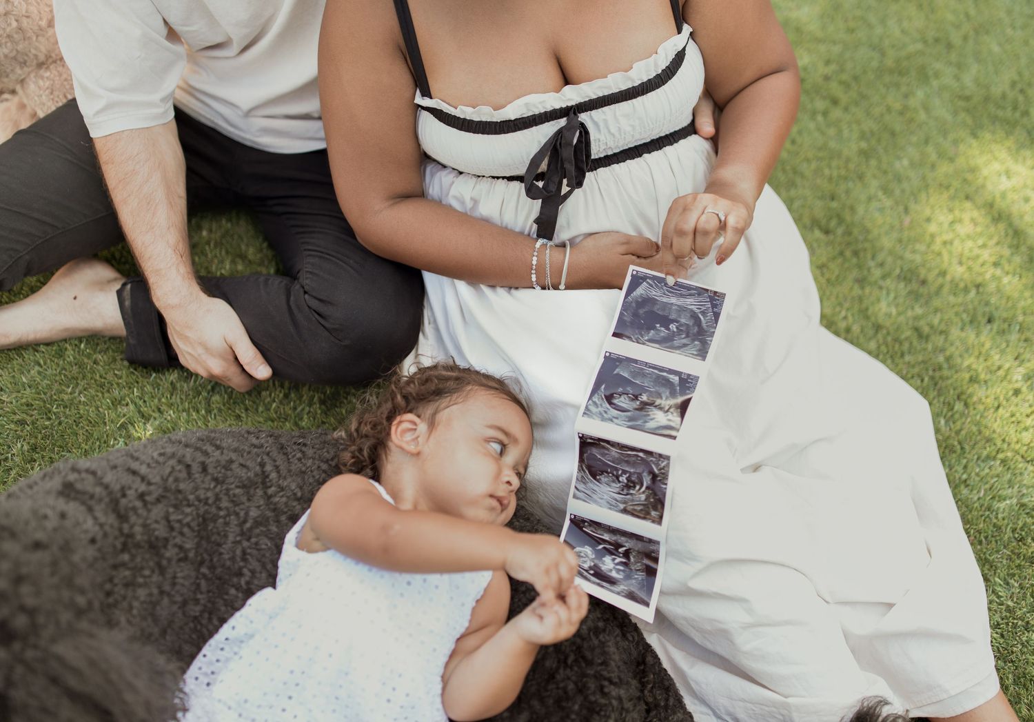 Expectant mother in a white dress holds ultrasound photos while a little girl lies on a blanket in the grass beside her.