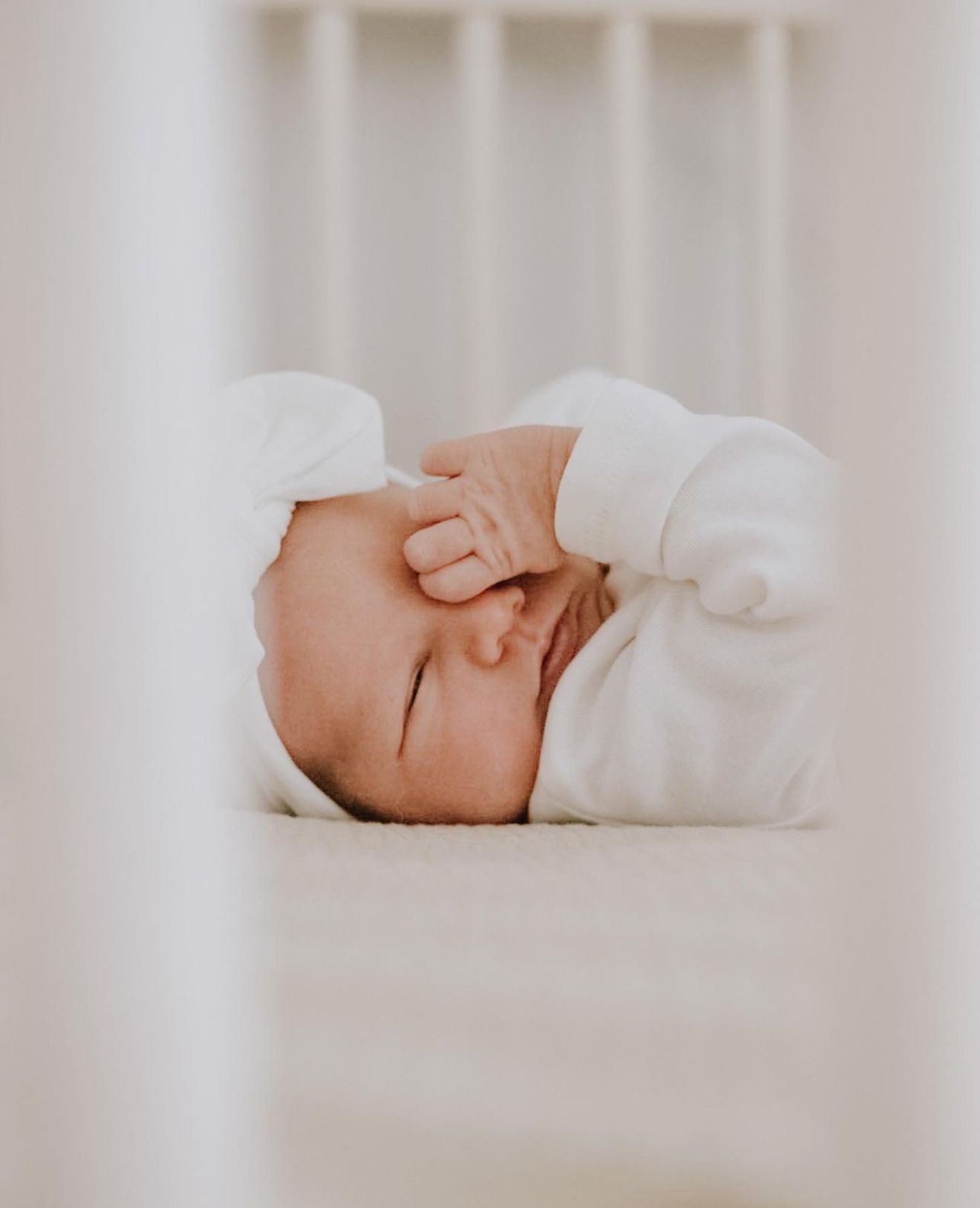 A newborn baby wrapped in a white blanket rubs their eyes while resting in a crib.
