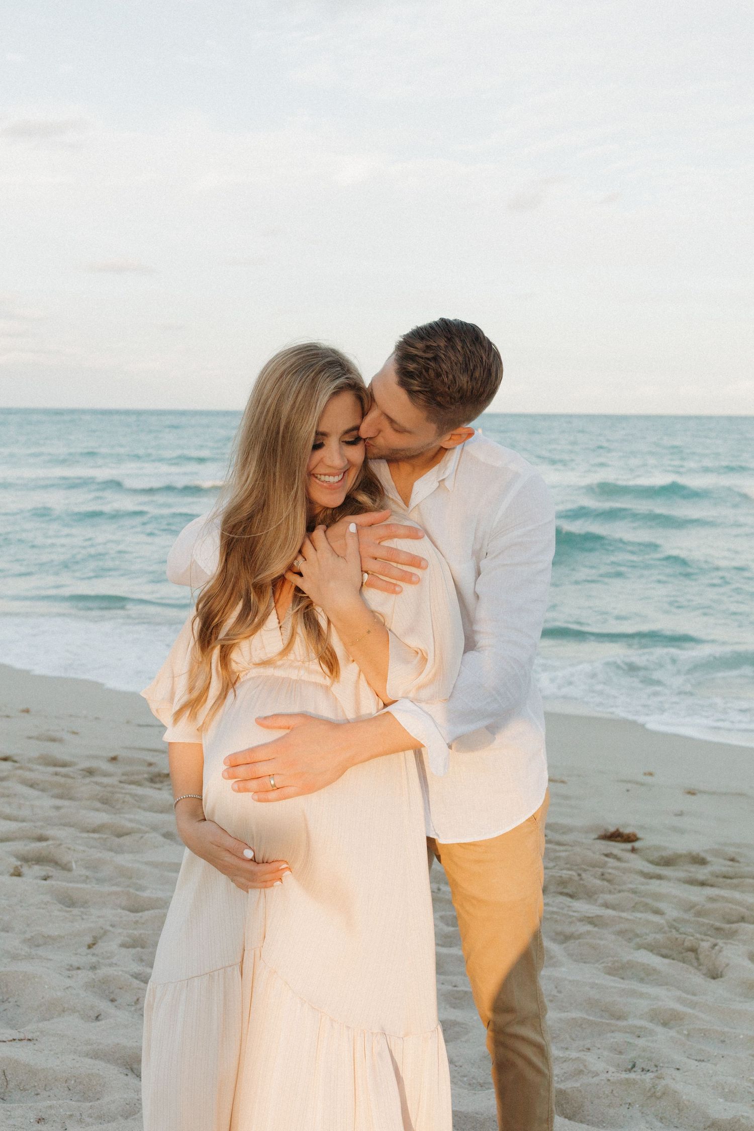 Expectant couple in cream and white outfits embrace on a sandy beach at sunset as he kisses her cheek and they cradle her baby bump.