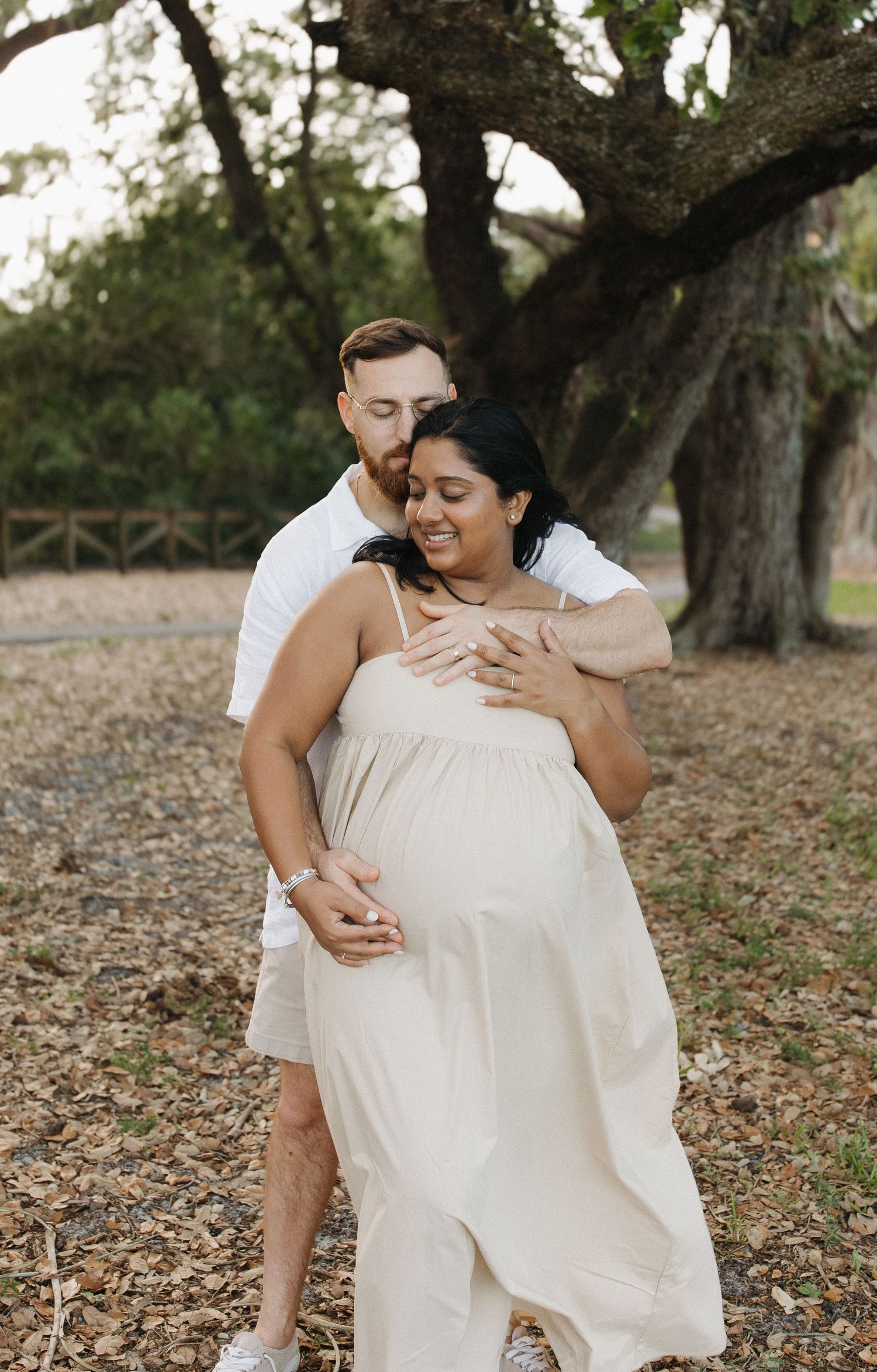 Smiling pregnant woman in a cream dress embraced from behind by her partner during a maternity photo shoot beneath a large oak tree.