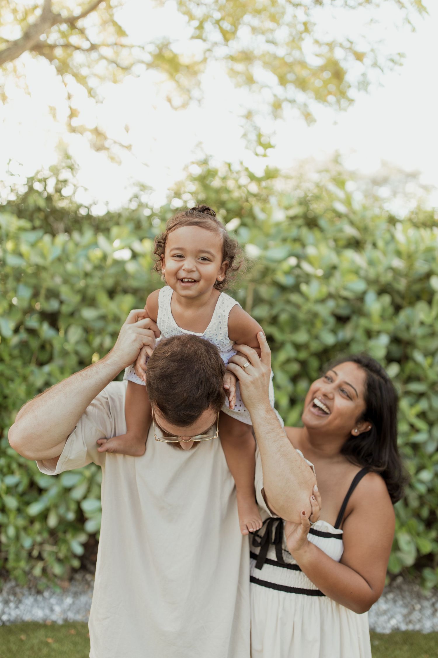 A laughing toddler sits on a man's shoulders while a smiling woman stands beside them in front of a green hedge in a joyful family portrait.