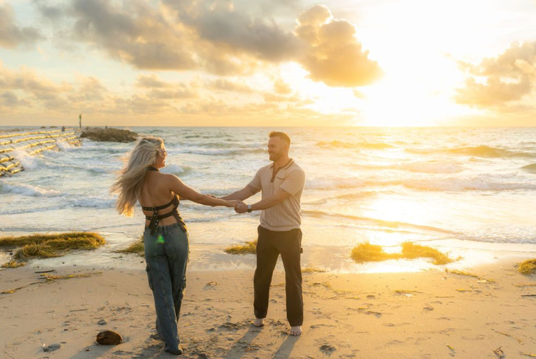 Two people dancing on a sandy beach at sunset with waves crashing against rocks in the background.