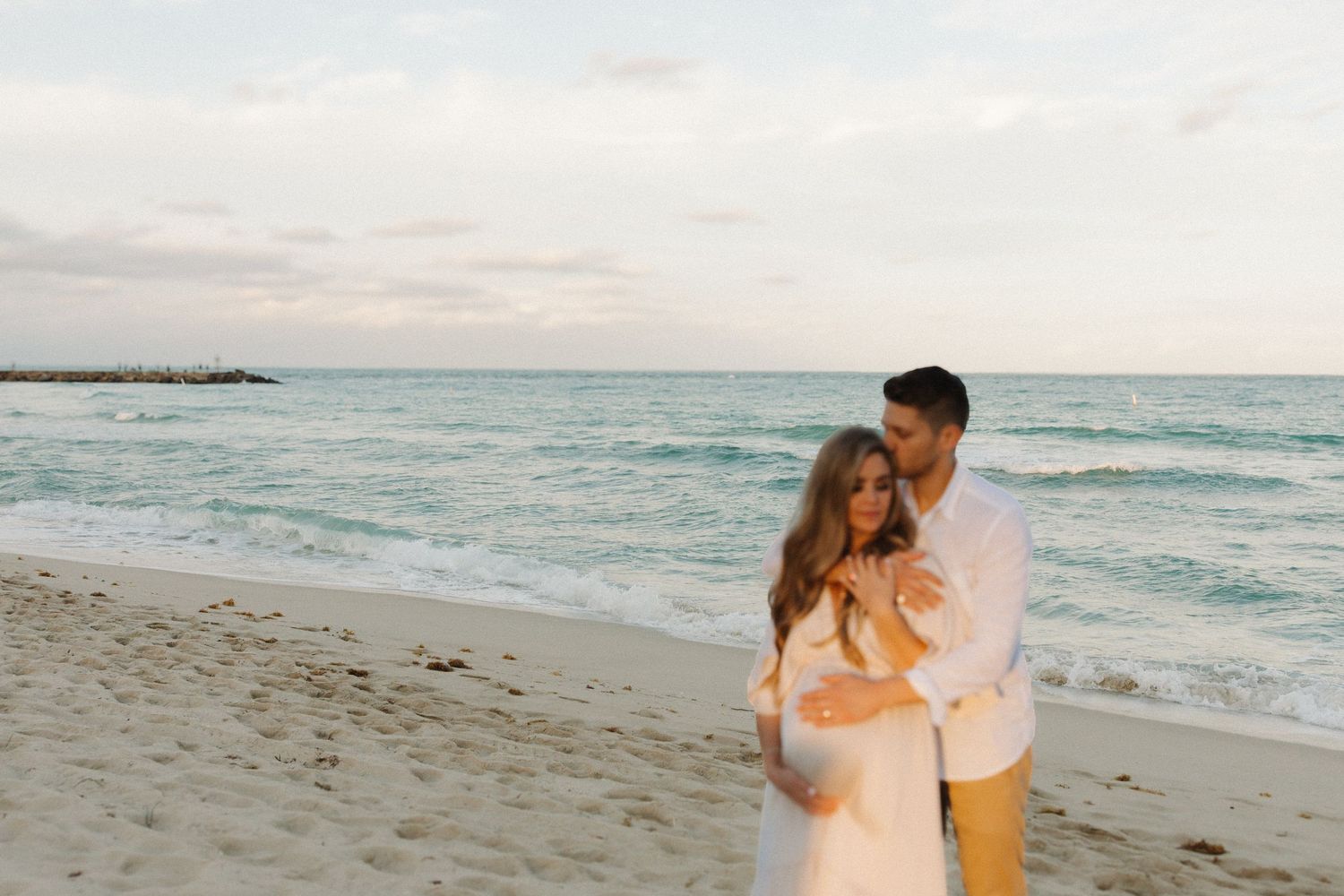 A couple embraces on a beach at sunset with turquoise waters and cloudy skies in the background.