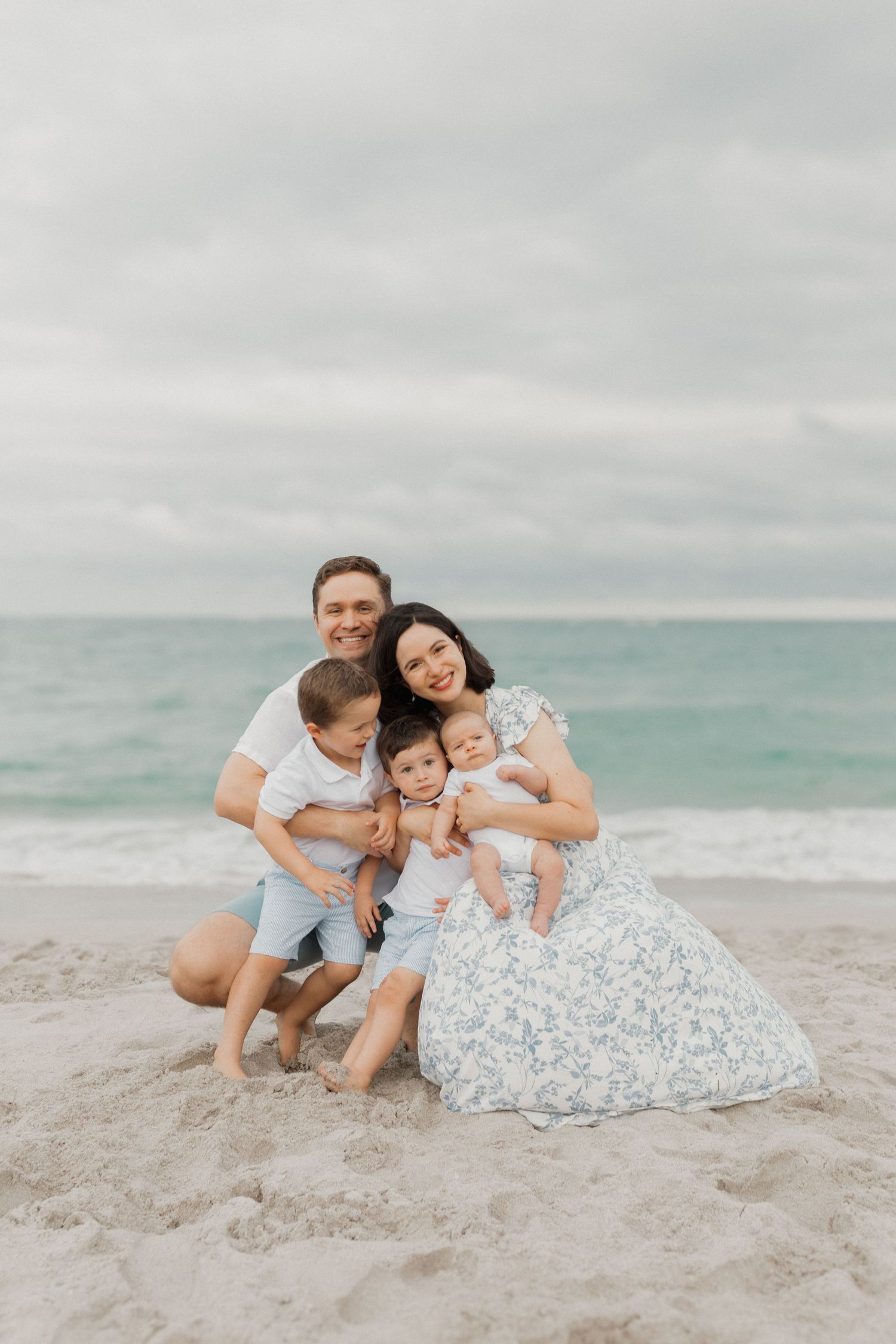 Family wearing white and blue outfits poses together on a cloudy beach with ocean in background.