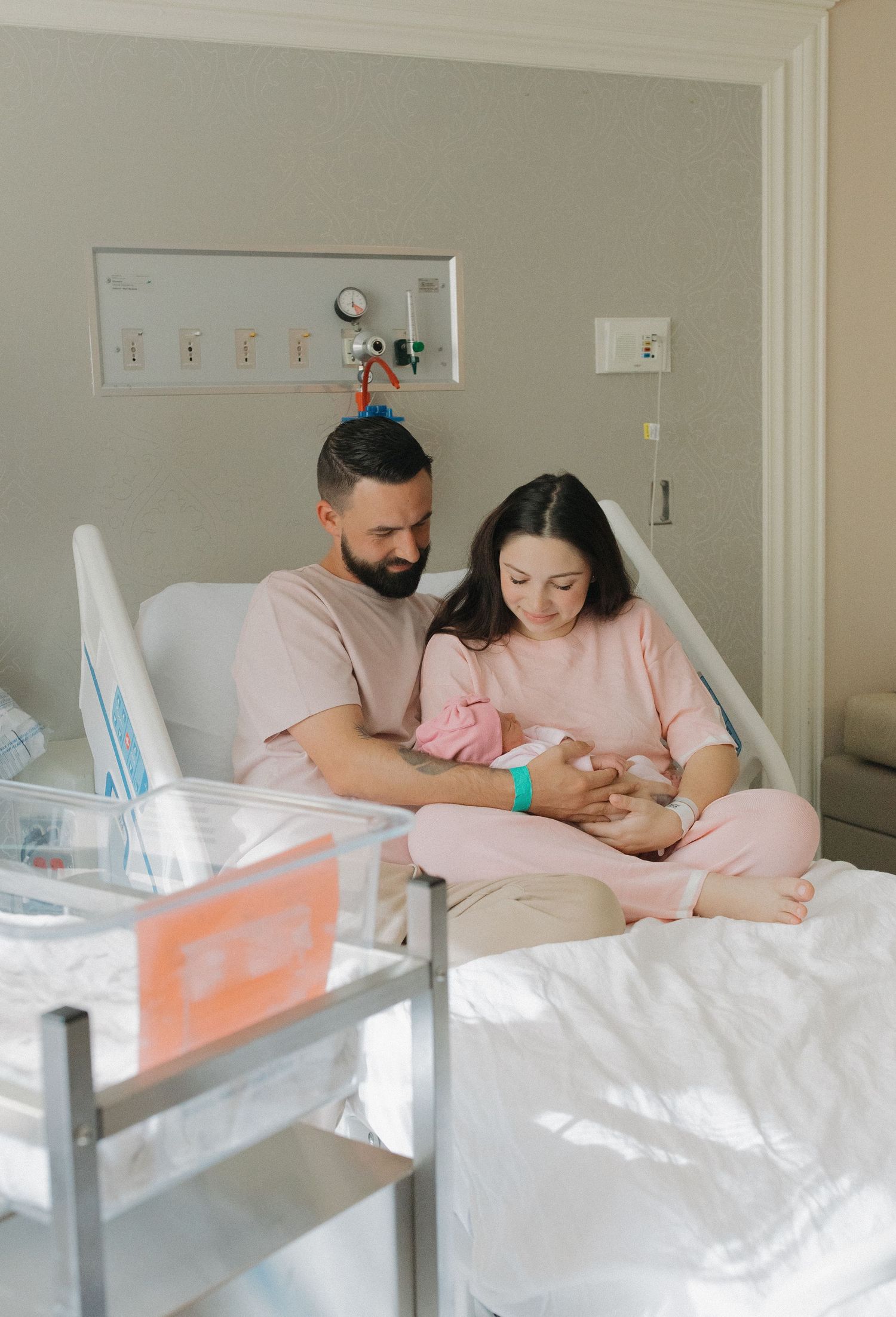 A couple in matching pink outfits sits together on a hospital bed while holding their newborn baby.