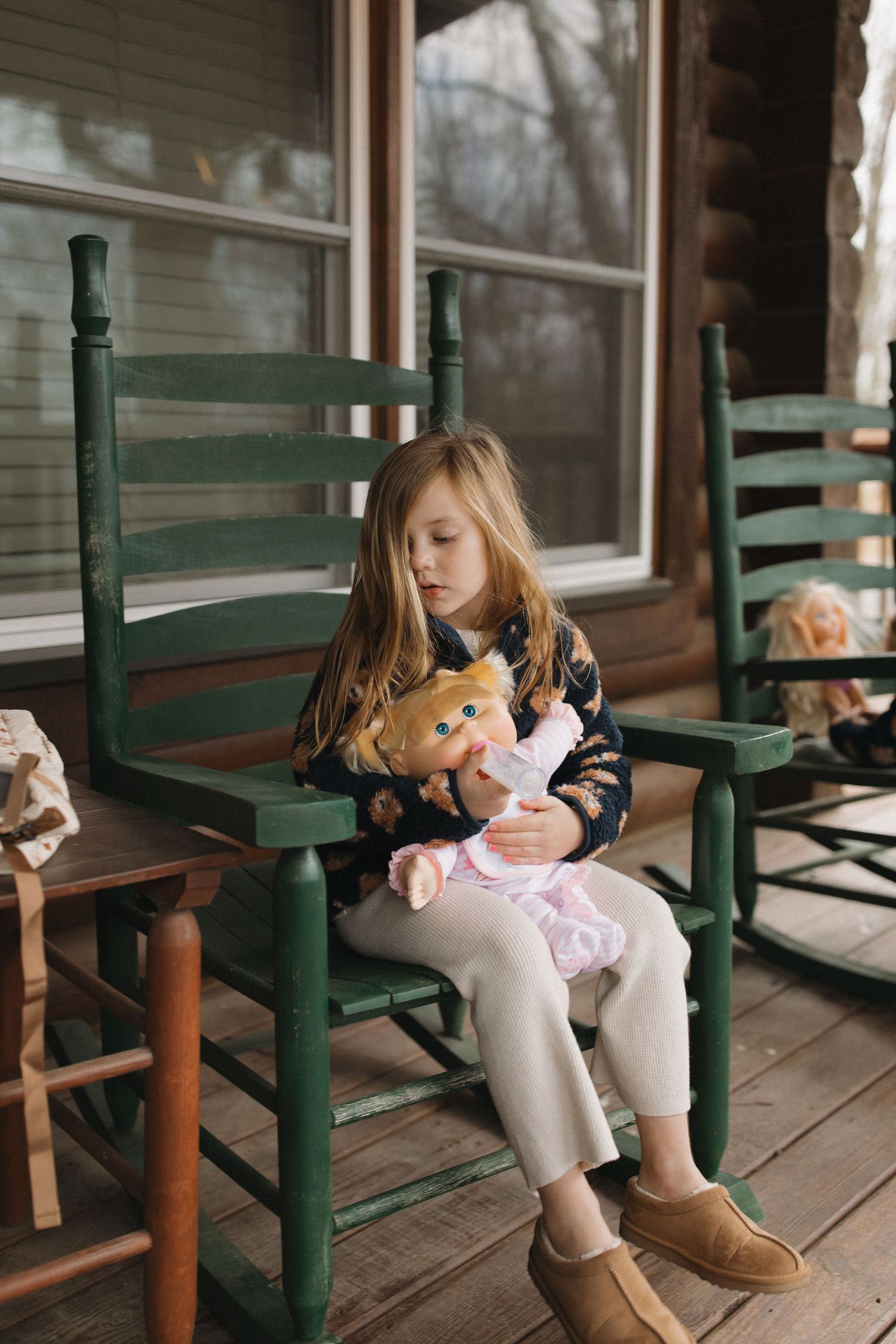 A child in a dark sweater and boots sits on a green wooden rocking chair on a porch holding a plush toy.