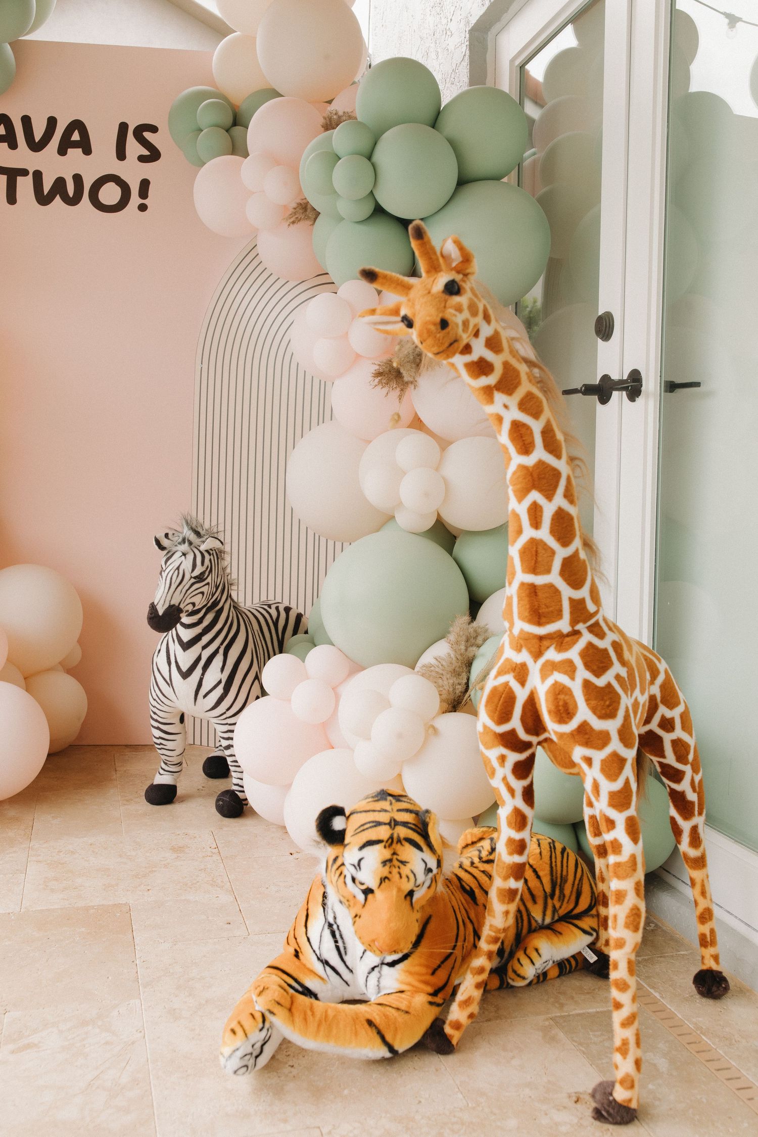 Safari themed party decoration with plush giraffe, tiger and zebra toys against pastel balloon arch in mint and peach colors.