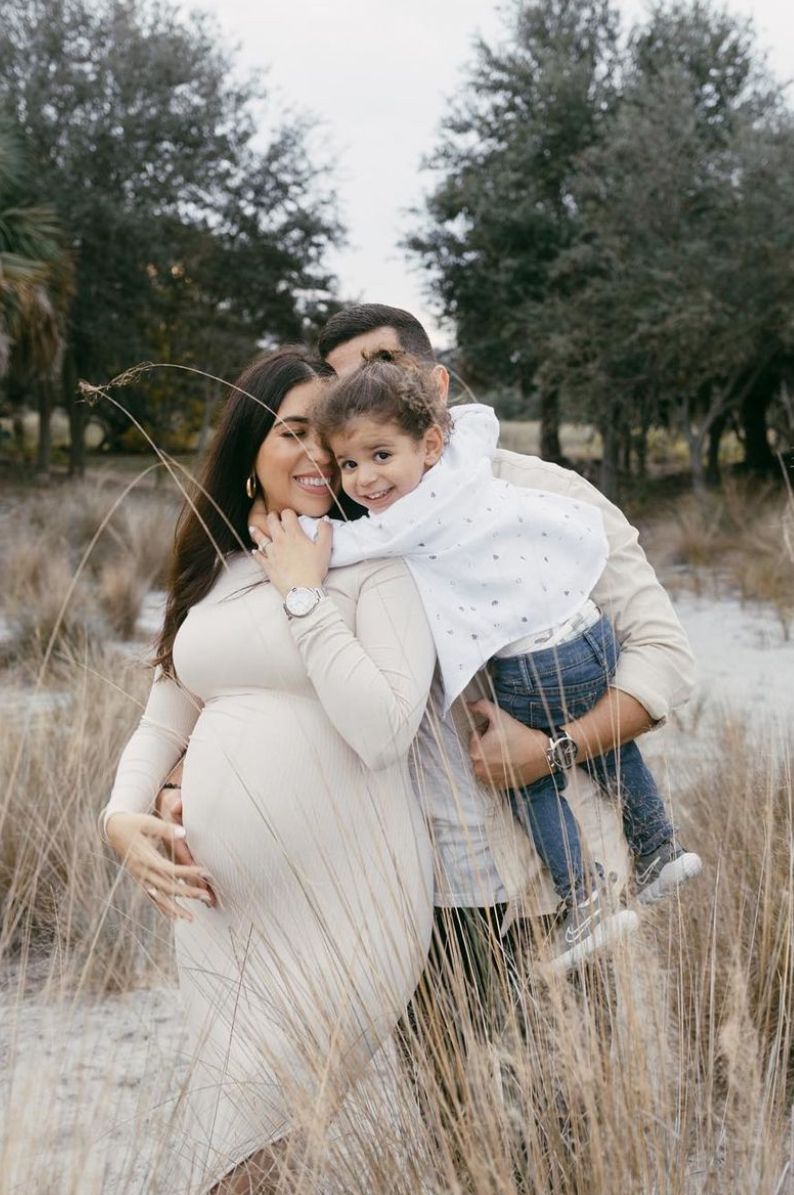 A family stands together in a field of tall winter grass, with the mother wearing a cream dress and cradling her baby bump.