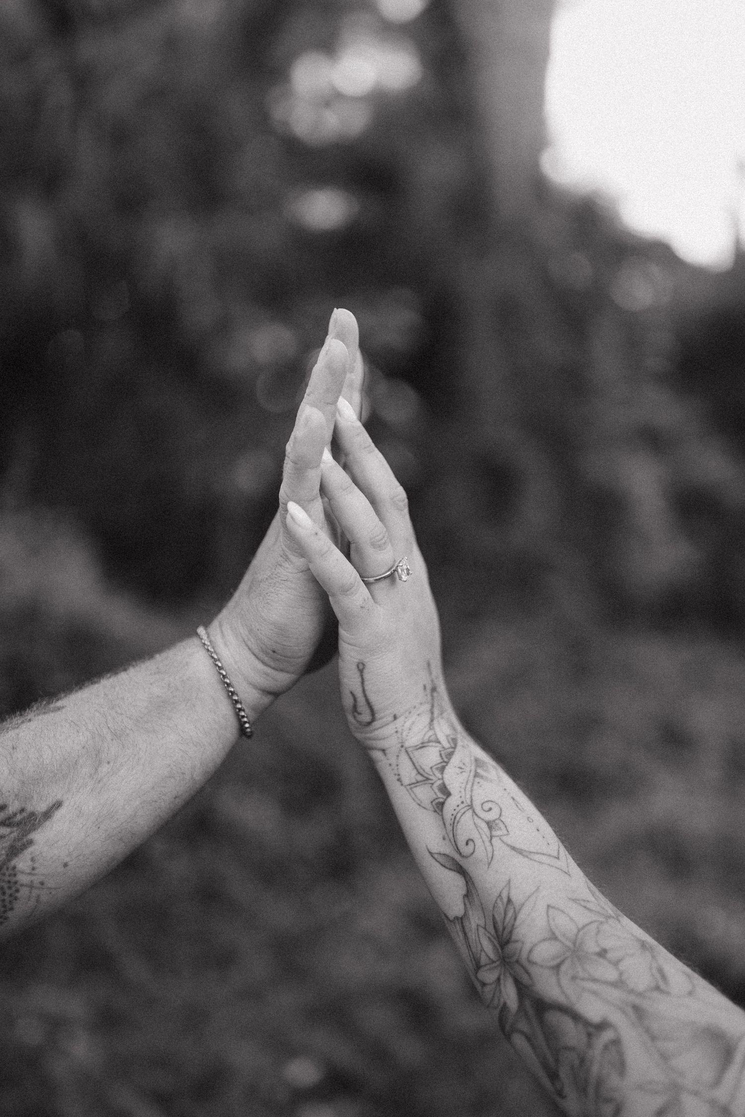Two hands with floral tattoos touch palm to palm in a black and white outdoor photo.