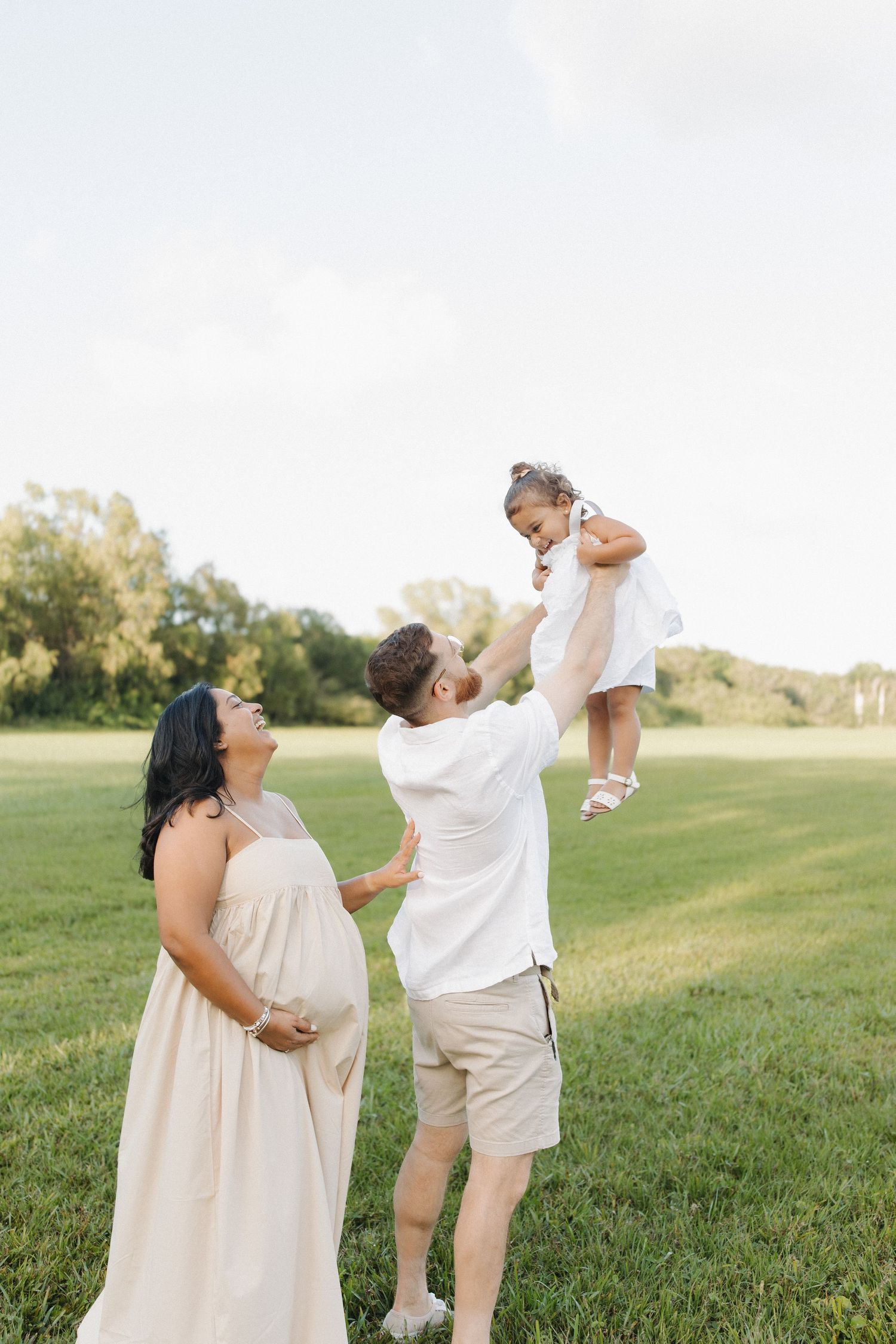 An expectant woman in a flowing white dress stands in a grassy field with a small child being lifted up high.