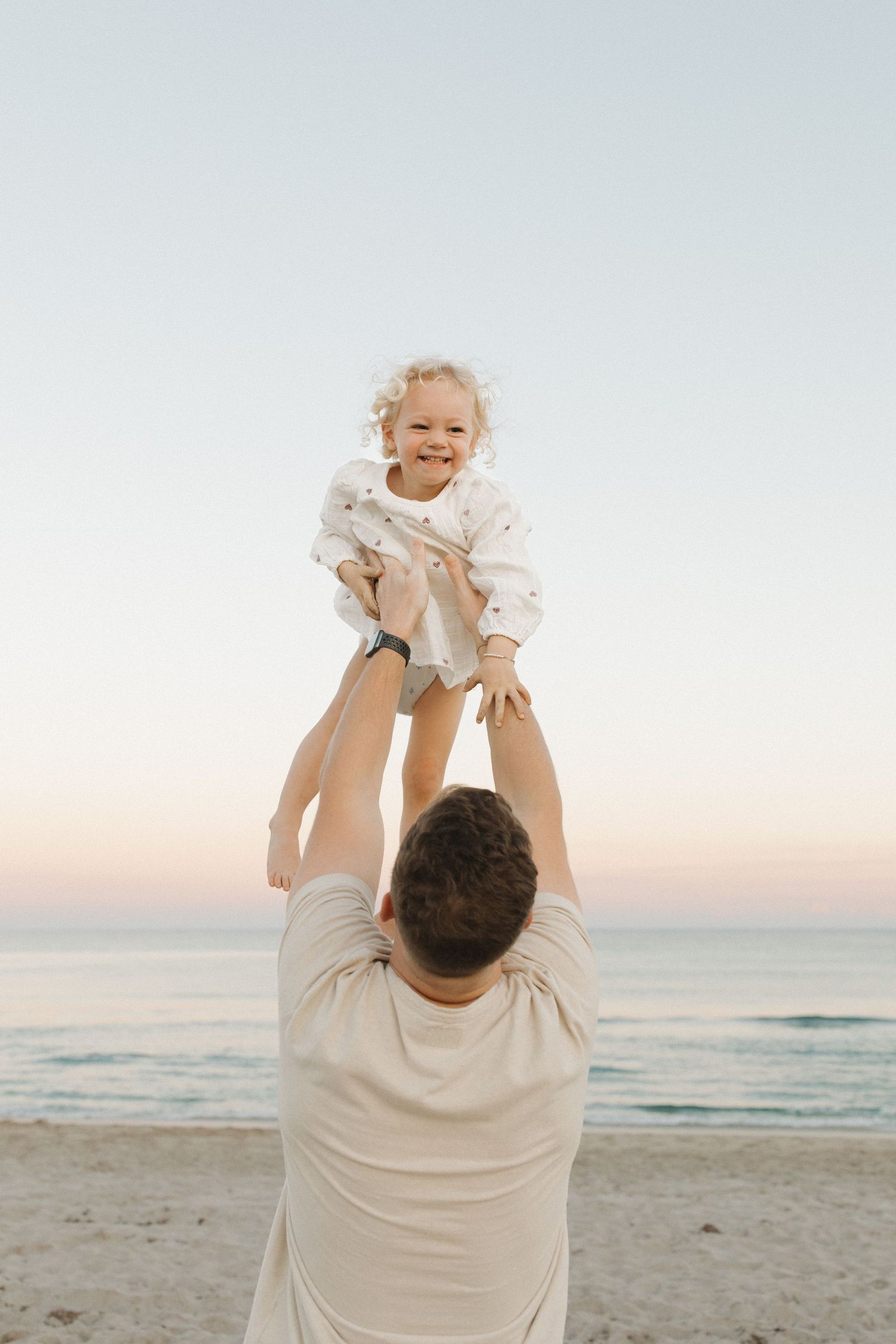 Father lifts a laughing curly-haired toddler in a white outfit high above him on a sandy beach at sunset.