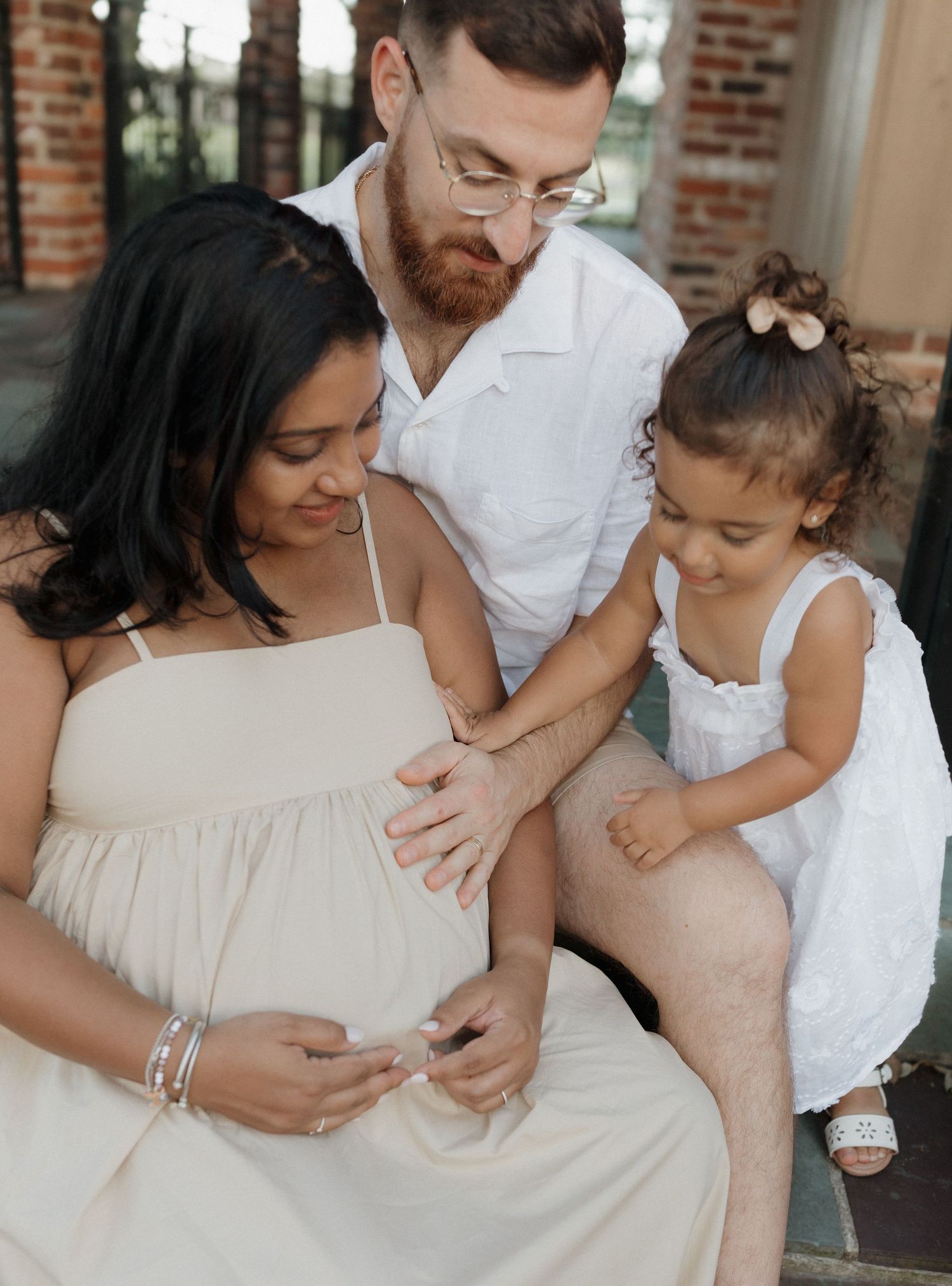 A family tenderly shares an intimate moment on their porch, touching a pregnant belly in a white maternity dress.