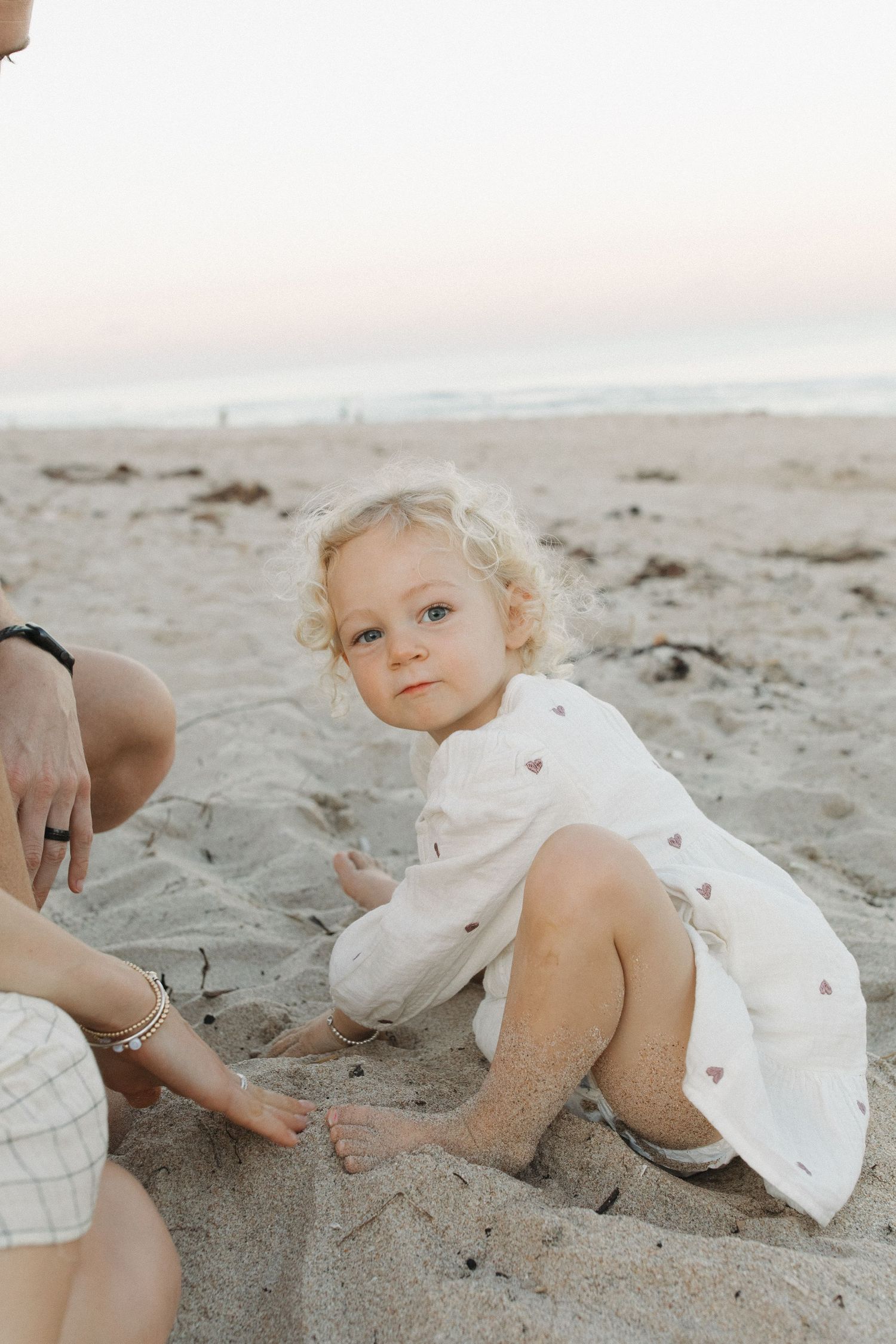 Young child wearing white dress plays in sand at beach during sunset with soft pink sky.