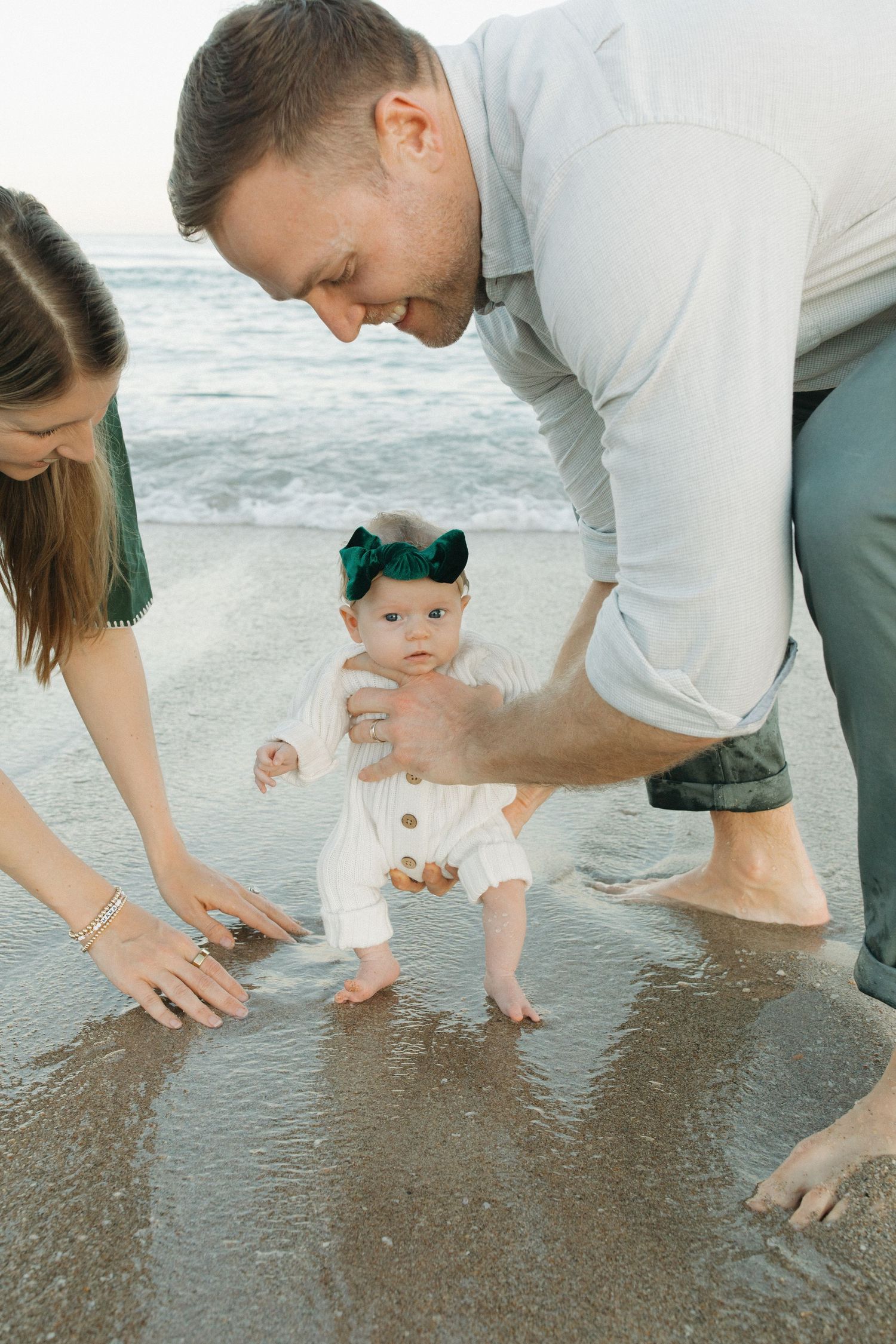 Parents help baby wearing turquoise bow take first steps on beach at sunset by the ocean waves.