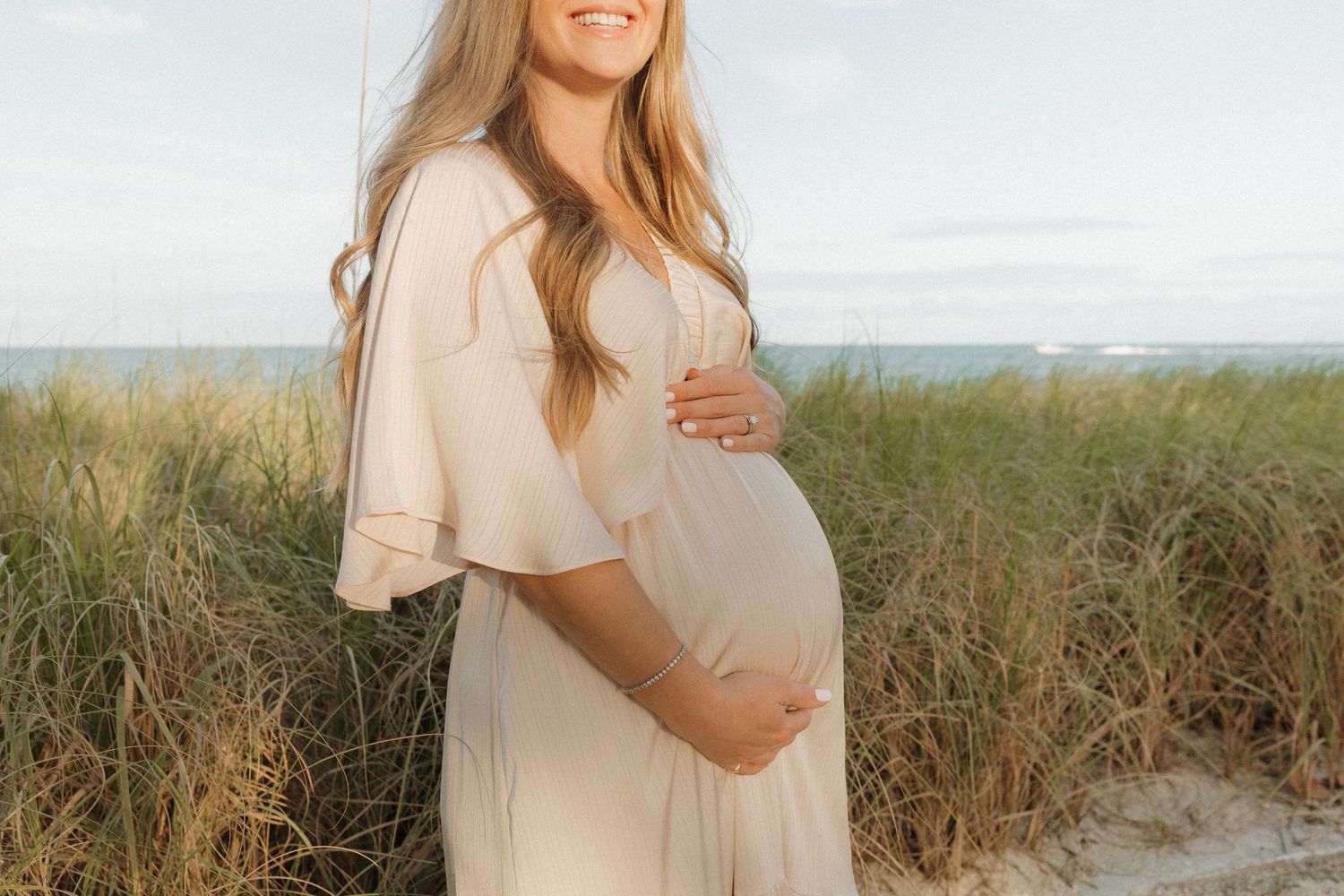 Smiling pregnant woman in a flowing cream dress cradling her baby bump while standing among beach grass and sand dunes by the ocean.