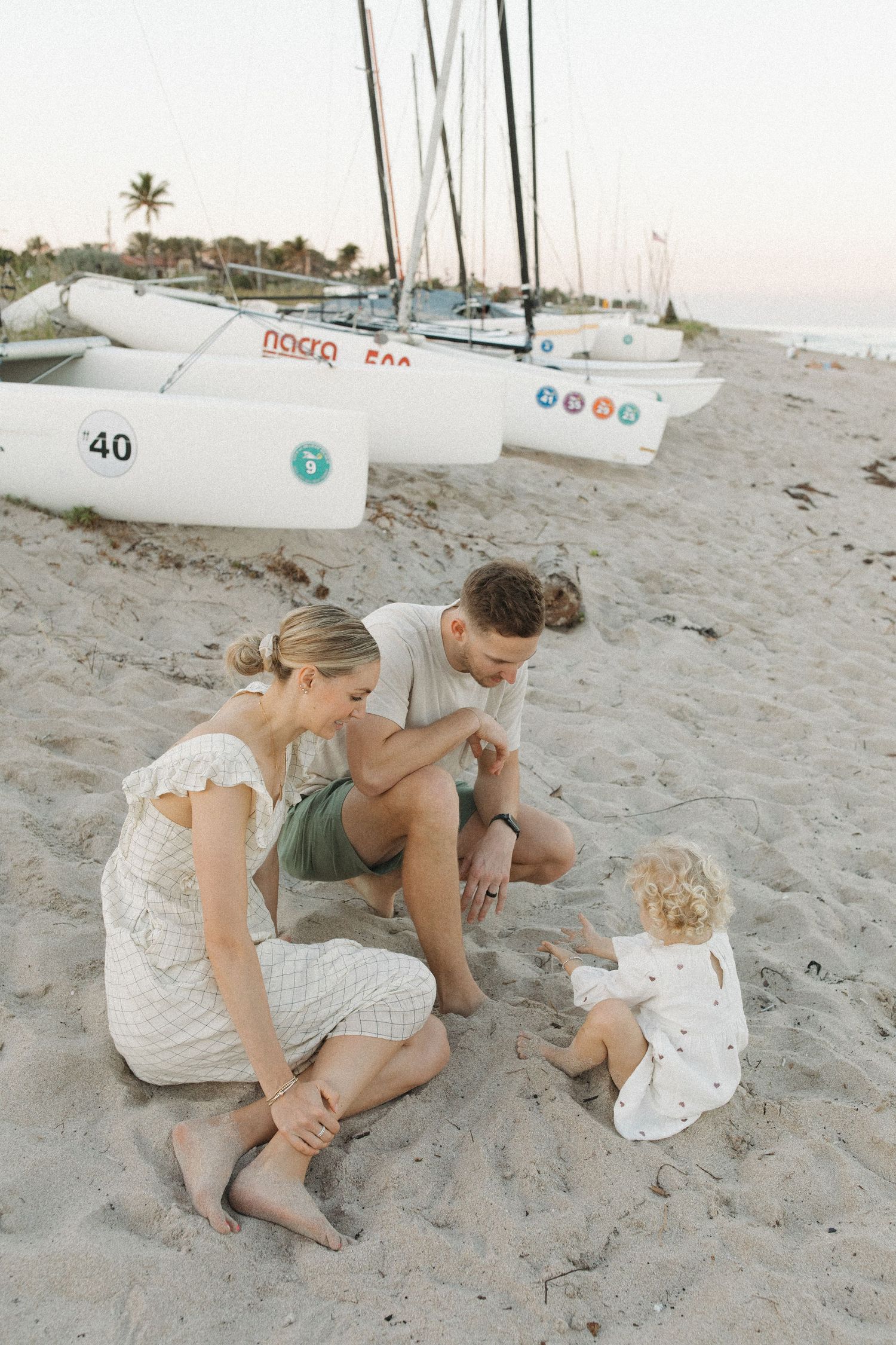 A young family of three sits together on a sandy beach at dusk, playing with their toddler near parked sailboats and palm trees.