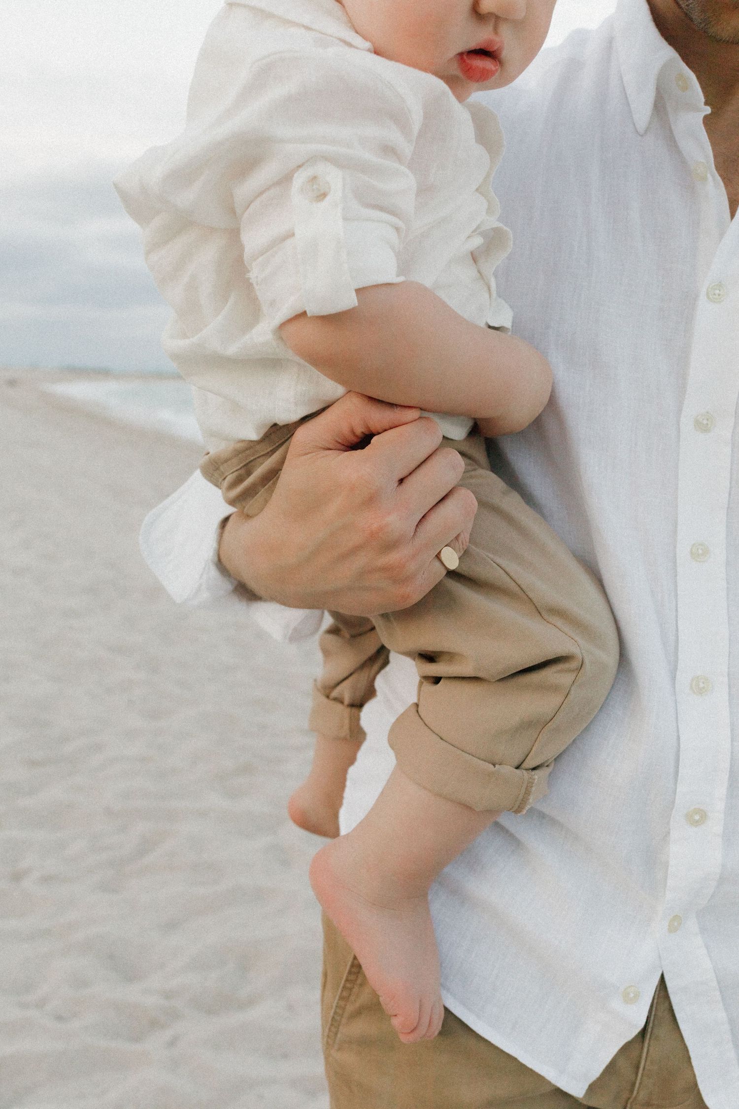 Adult in a white linen shirt and khaki pants holds a barefoot baby in a matching outfit on a sandy beach.