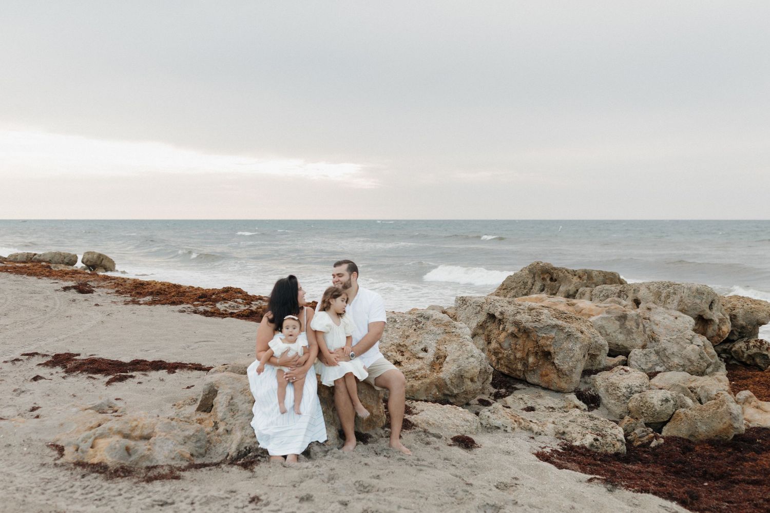 Family of three in white outfits sits together on large rocks along a sandy beach with ocean waves and a soft sunset sky behind them.